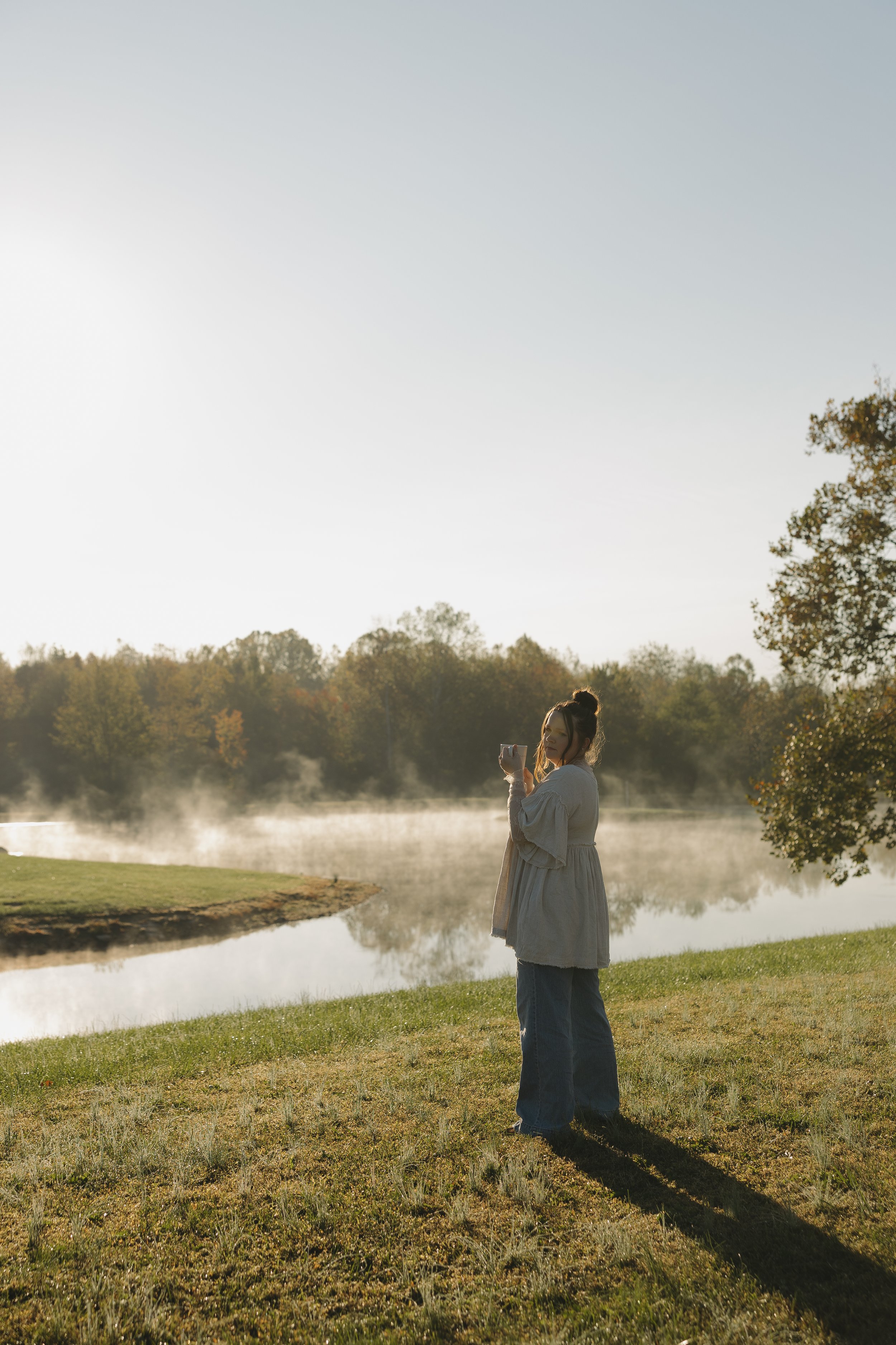 A woman in a long coat and jeans standing on grass near a river or lake, holding a mug, with trees and mist in the background during sunrise or sunset.