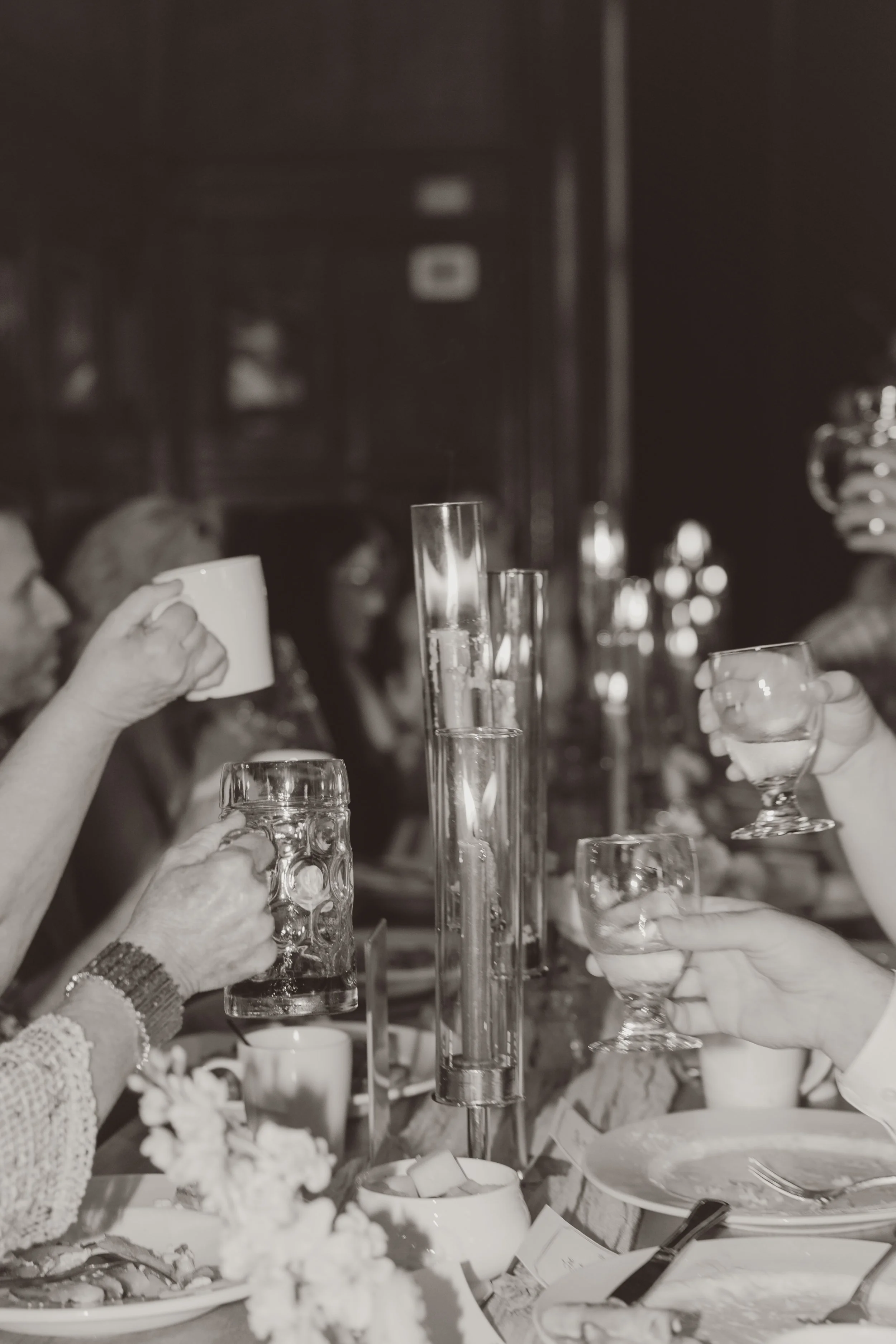 People raising glasses in a toast at a dining table with various glasses and dishes, in a dimly lit restaurant or cafe.