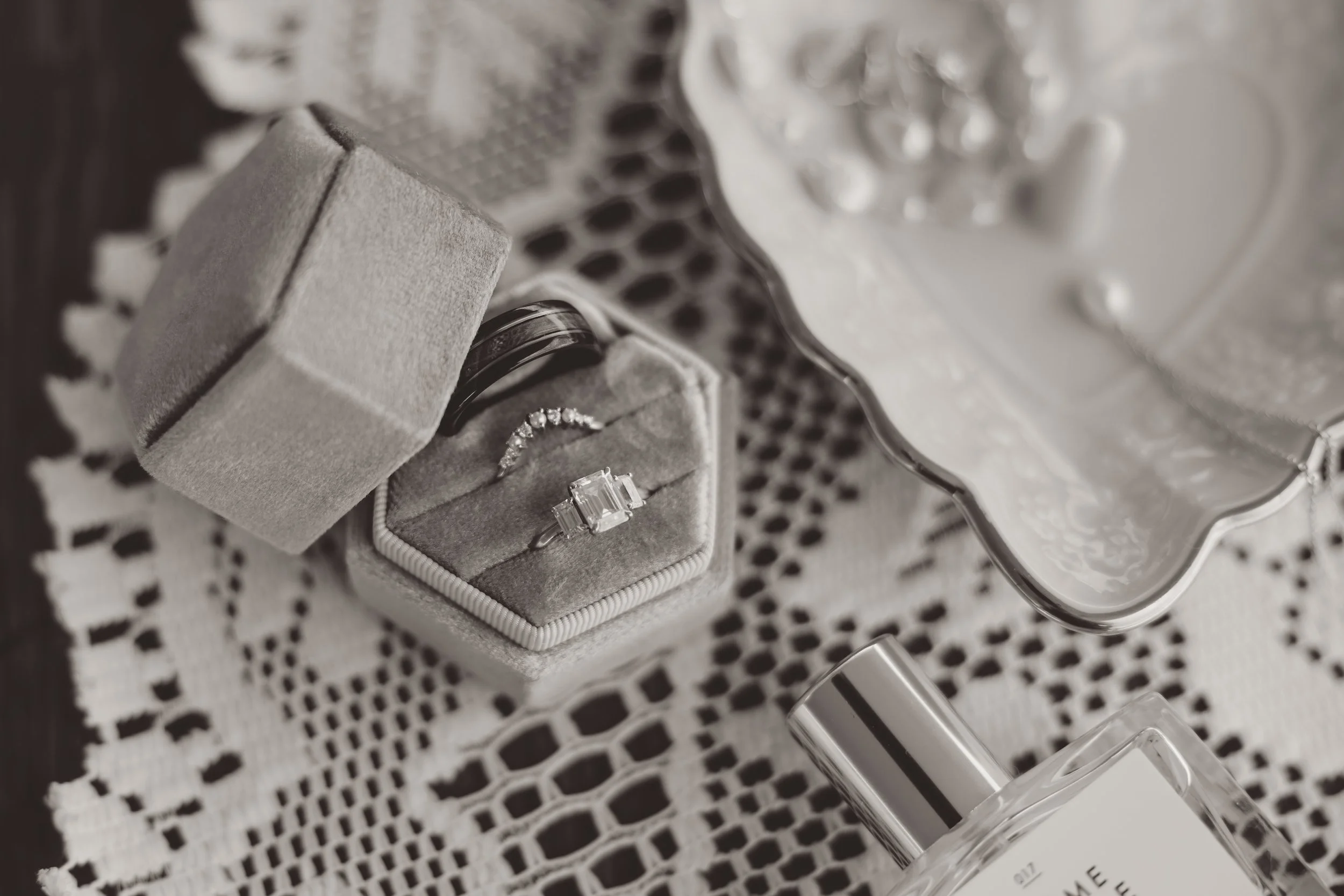 Close-up of jewelry in a velvet ring box, including an engagement ring with diamonds, resting on a lace cloth along with a perfume bottle and a decorative dish.