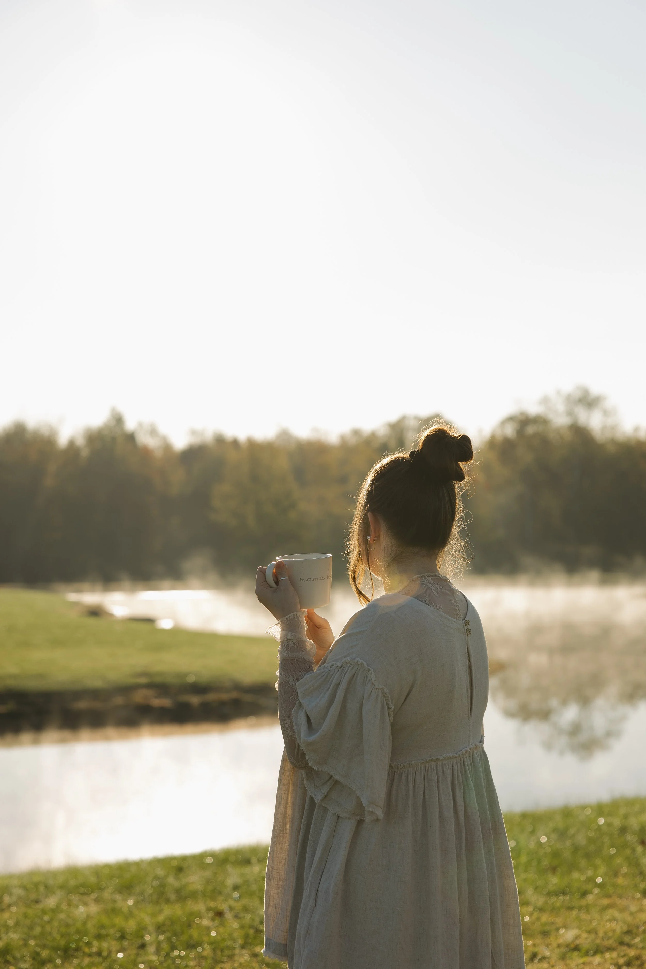 A woman in a light dress holding a mug and looking at a peaceful lake reflecting the trees in the background during sunrise or sunset.