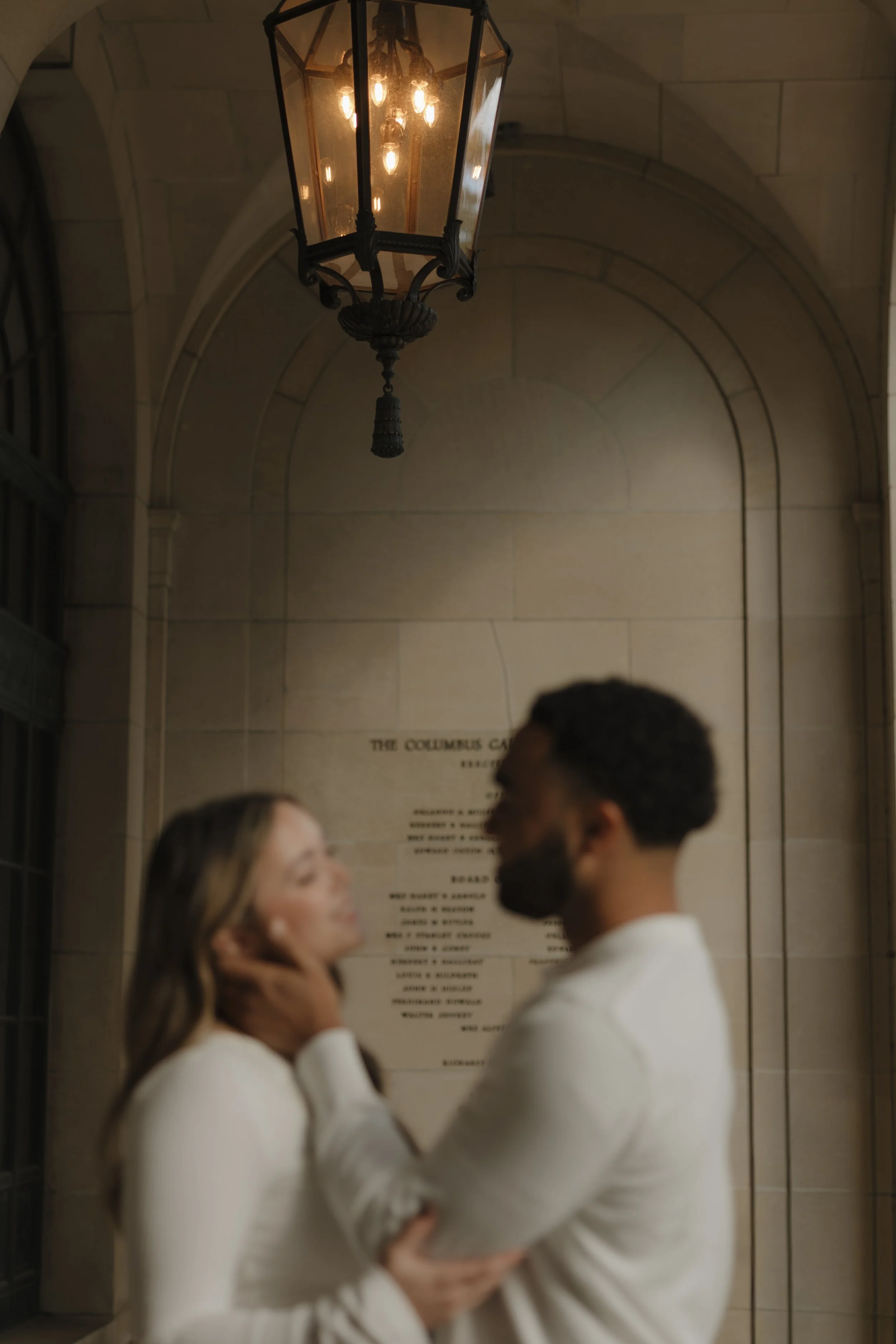 A couple standing close together indoors, with a lantern hanging above them and a plaque behind, engaging in an intimate moment.