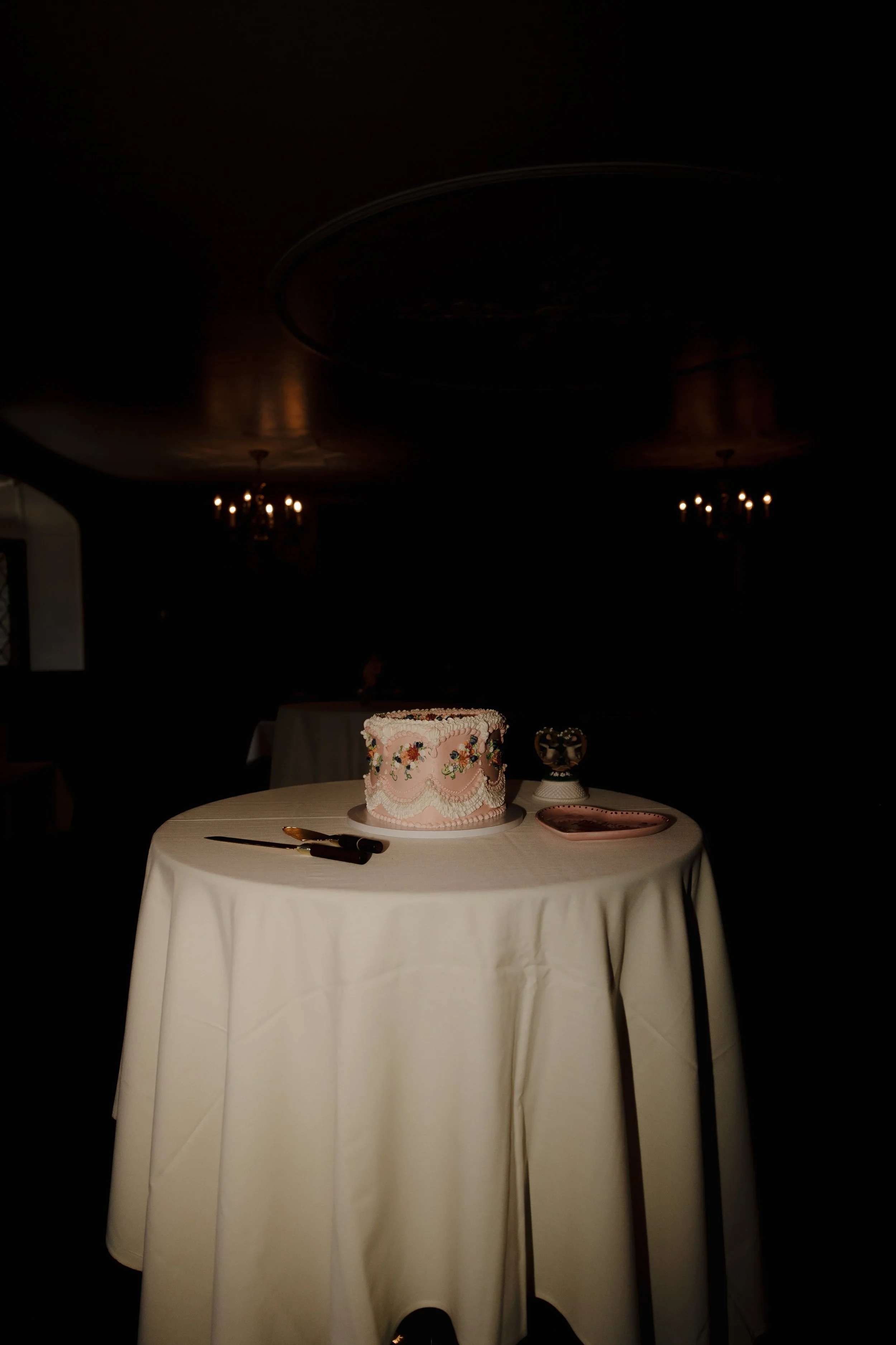 A round table with a white tablecloth holding a decorated cake, a knife, a pink plate, and a small bowl, in a dimly lit room with chandeliers.