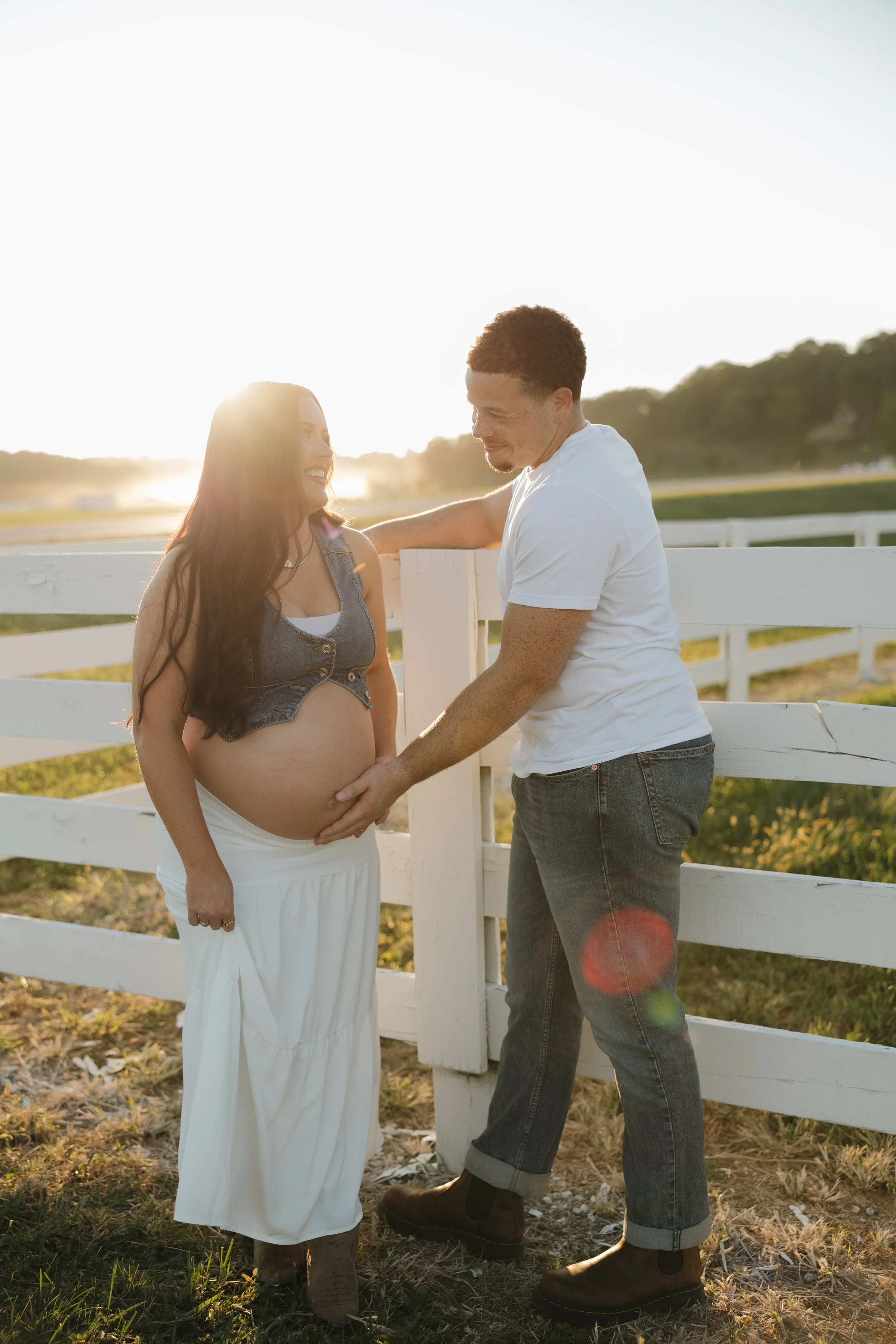 A pregnant woman and a man standing near a white fence in a field at sunset, with the man touching her belly and both smiling.