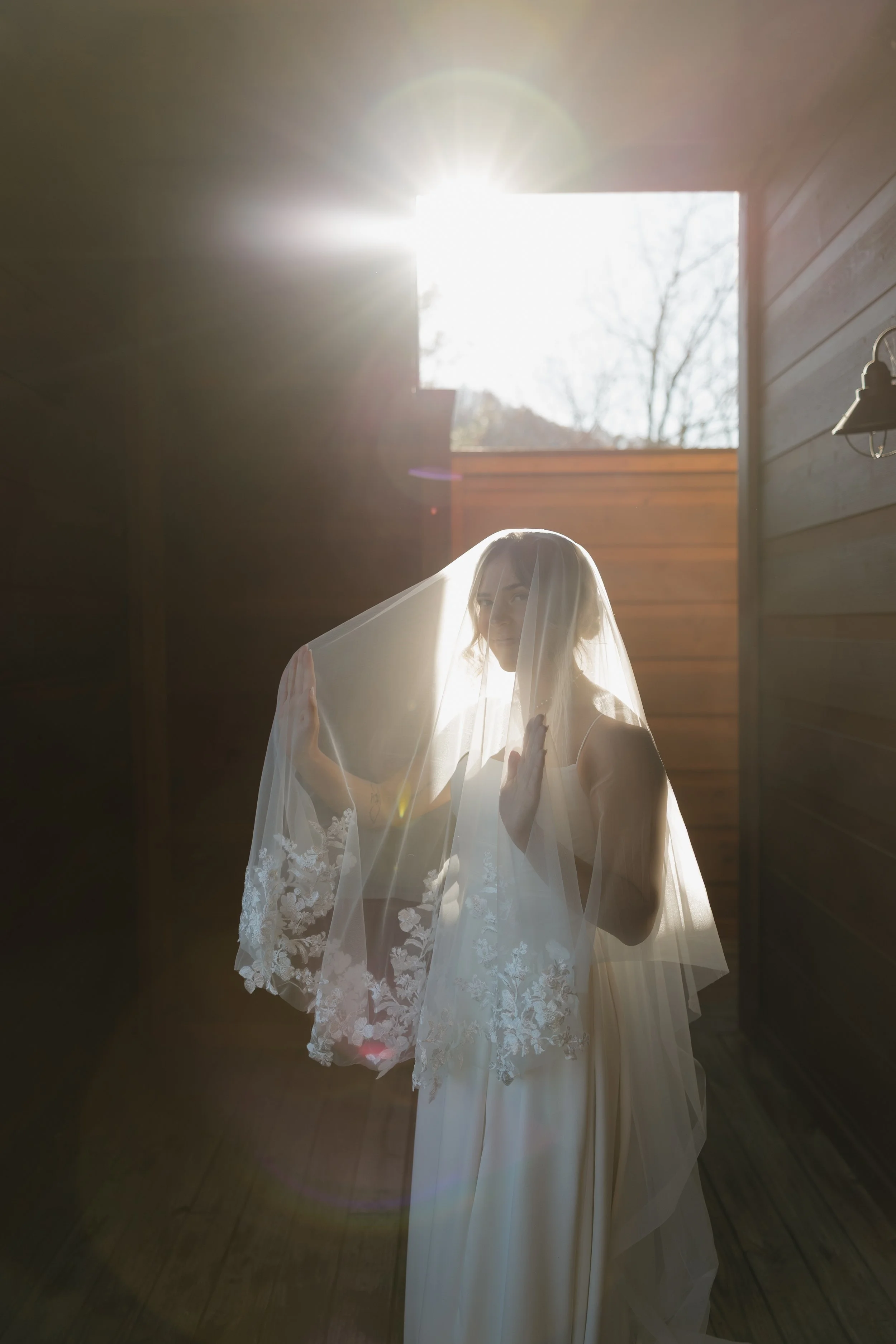 Bride standing indoors with sunlight shining through an open window behind her, wearing a wedding dress and veil, holding the veil with both hands and looking at the camera.