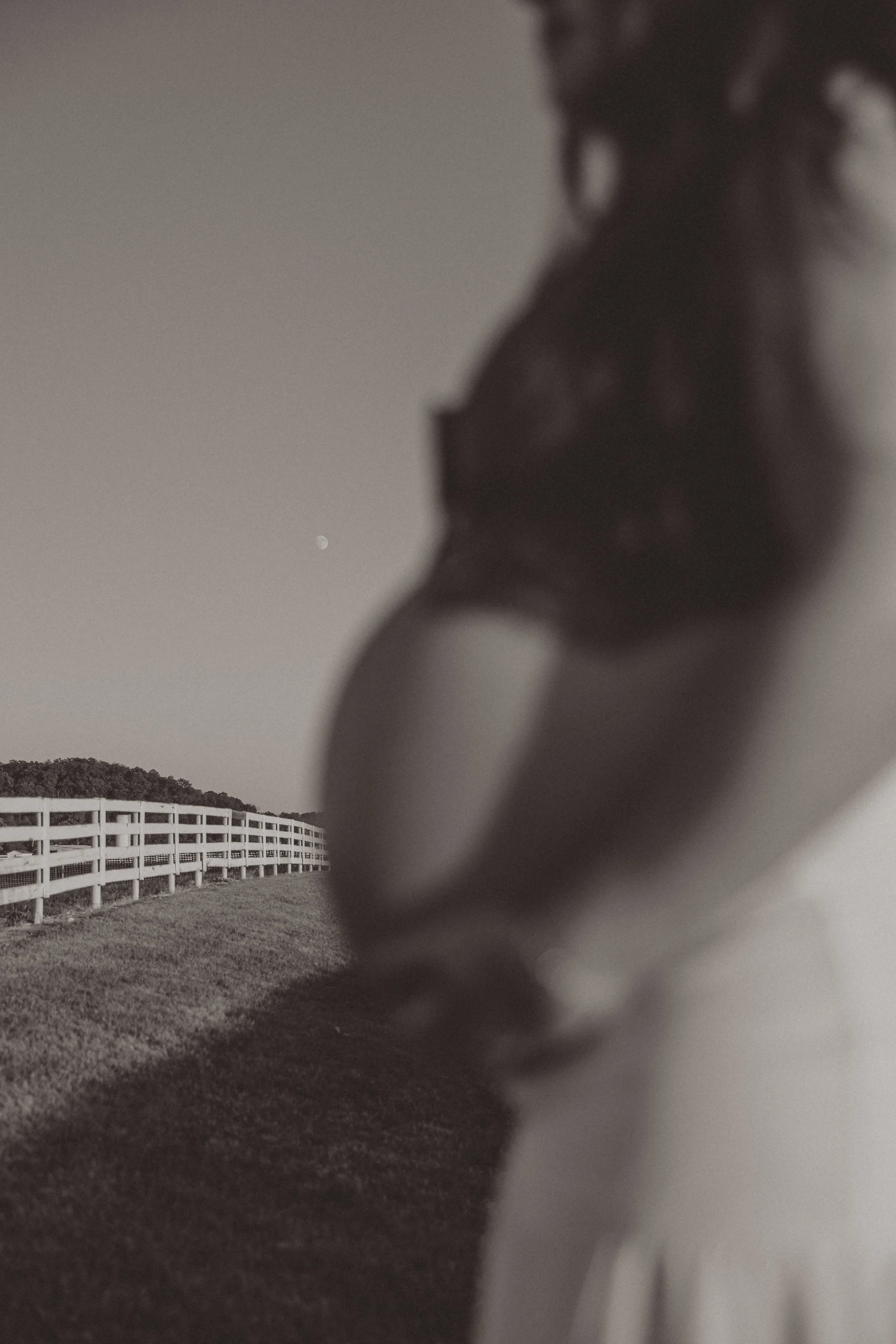 Blurred photo of a person standing outdoors near a white fence, with a distant hill and the moon in the sky.