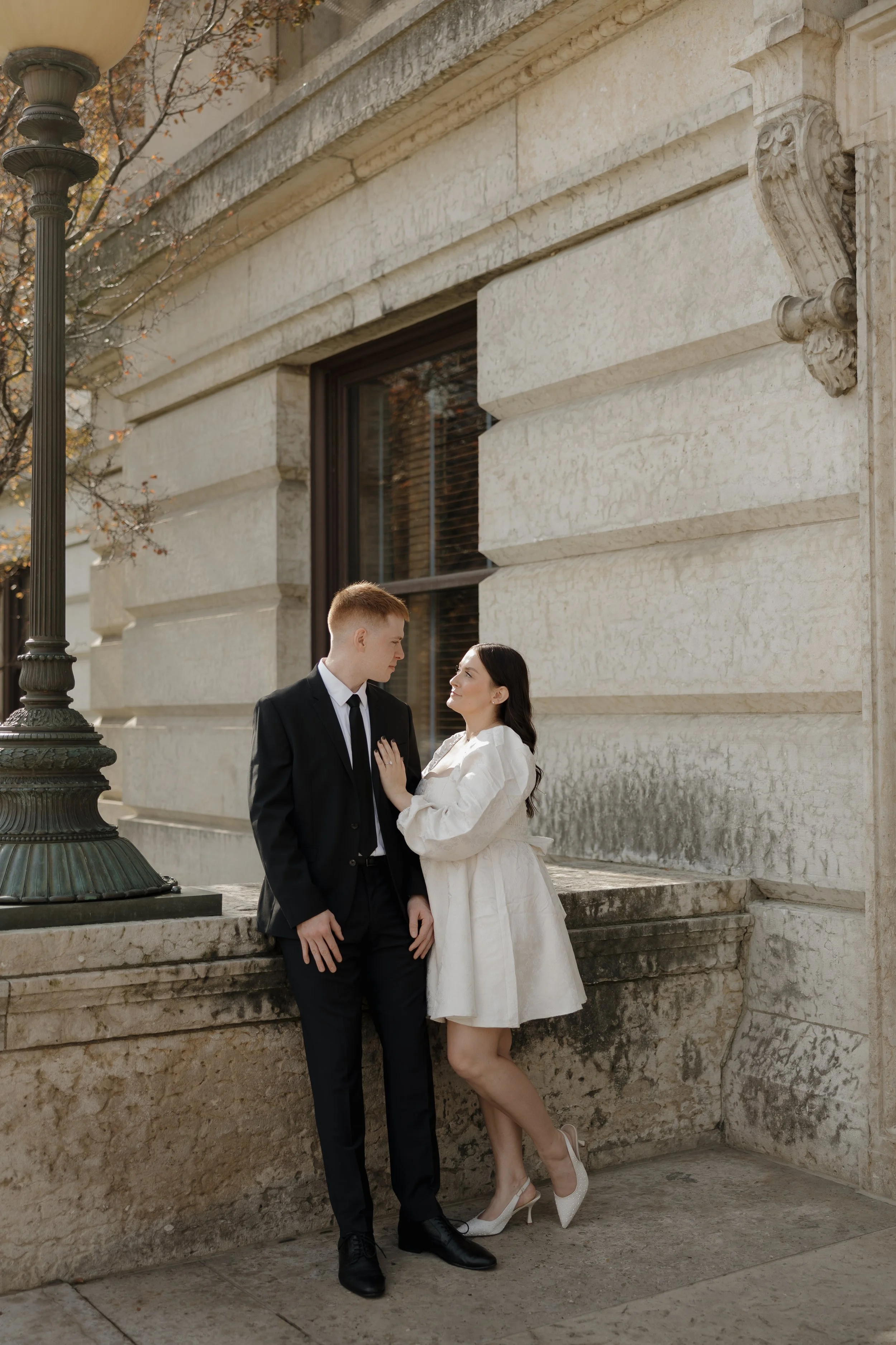 A couple dressed in formal attire standing close to each other outdoors near an ornate stone building. The man is in a black suit, and the woman is in a white dress and white high heels.