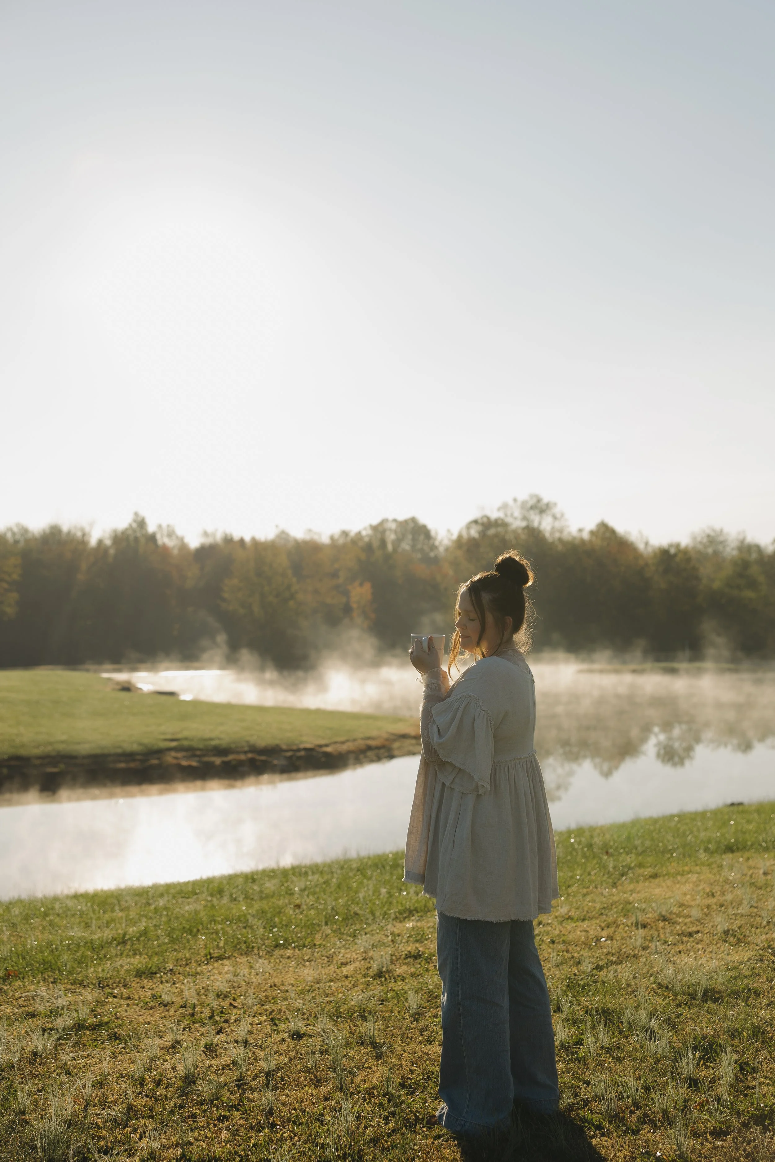 A woman standing near a body of water, holding a mug, with steam rising from the water. She is wearing a cream-colored dress and blue jeans, with her hair in a bun, during sunrise or sunset.