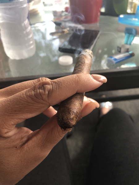 A person holding a lit cigar with smoke rising from it, sitting at a table with various items including a water bottle, smartphone, and medication.