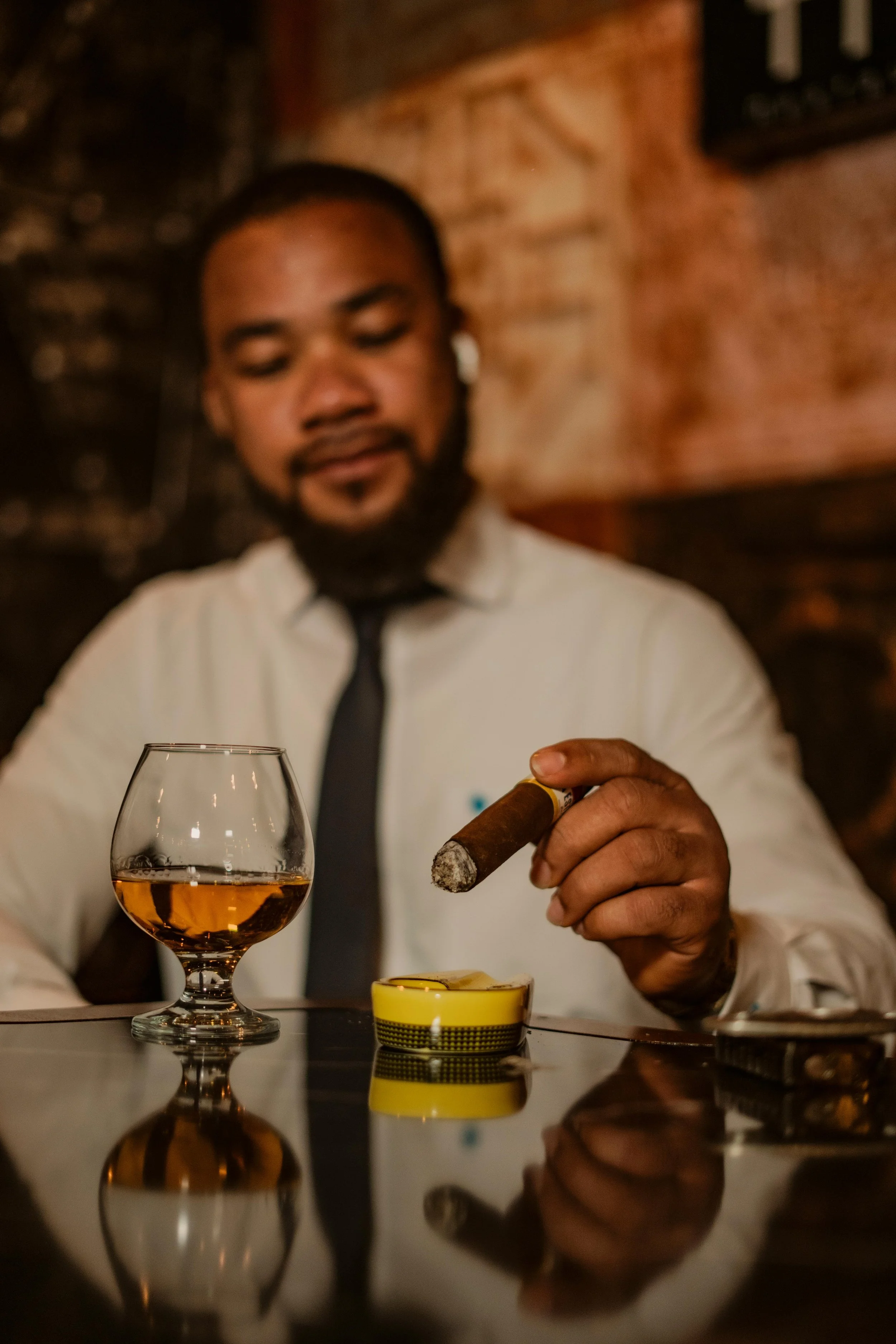 A man dressed in a white shirt and black tie sitting at a table with a glass of whiskey, holding a cigar, in a dimly lit bar setting.