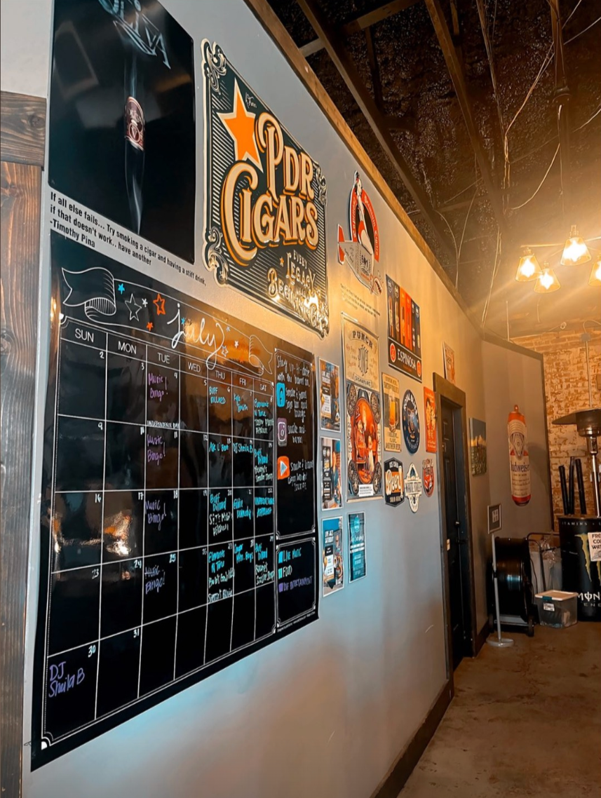 A wall in a bar or cafe decorated with various signs, posters, and a large monthly events calendar. The calendar shows events for July, including music bingo and DJ schedules. The wall is illuminated by warm hanging lights and has an exposed ceiling with wooden beams and wires.