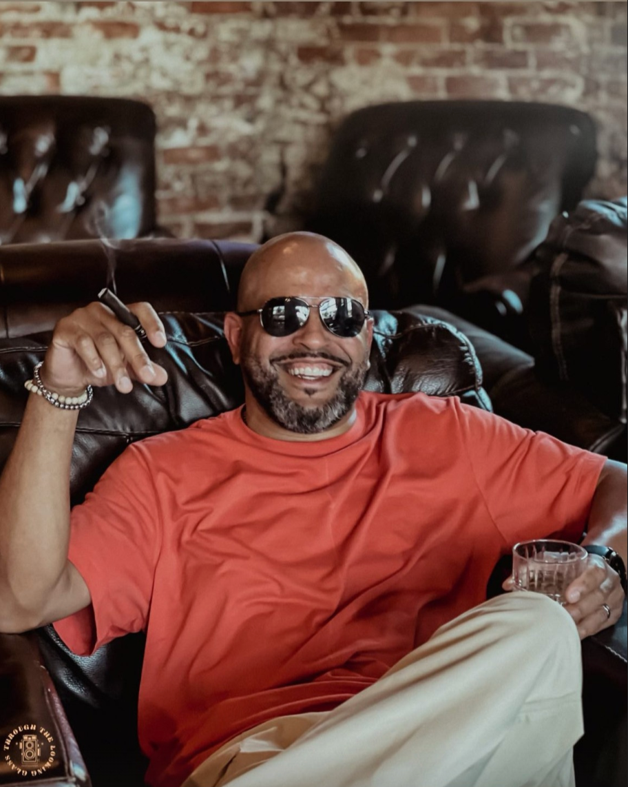 Man with a beard and sunglasses sitting on a leather couch, smiling, holding a cigar in one hand and a glass in the other, wearing a red shirt and beige pants, in a cozy, brick-walled setting.