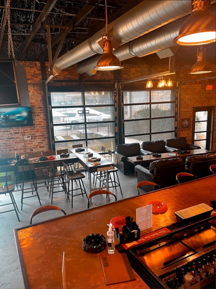 Interior of a modern restaurant or bar with high tables, stools, and leather couches, exposed brick walls, large windows, industrial lighting fixtures, and a view of parked cars outside.
