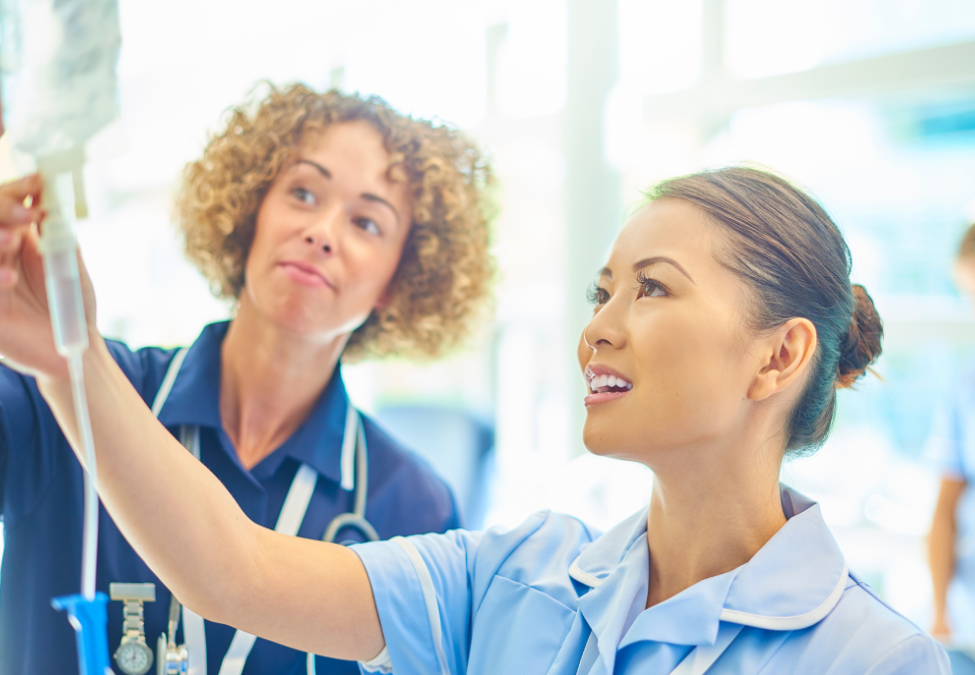 Two nurses examine a test tube in a brightly lit hospital or clinic setting.