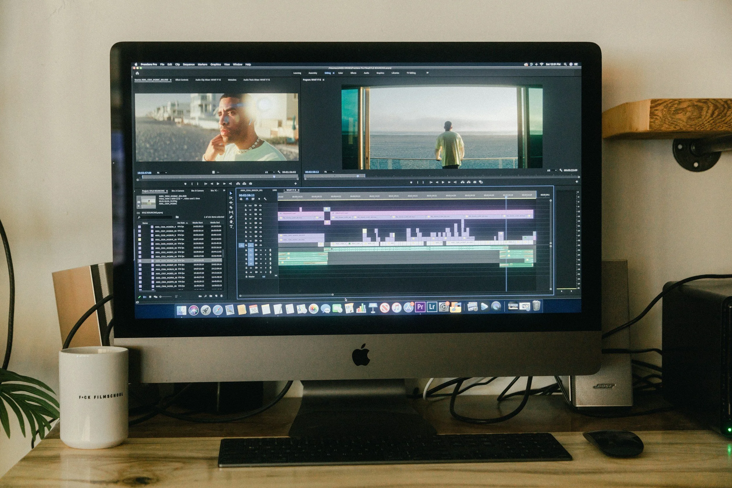 A computer monitor displaying video editing software with multiple video and audio tracks, with images of a person looking at the ocean on the screen. The monitor sits on a wooden desk with a keyboard, mouse, a white cup labeled 'FCK FilmSchool,' and some cables, with a plant on the left and a small speaker on the right.