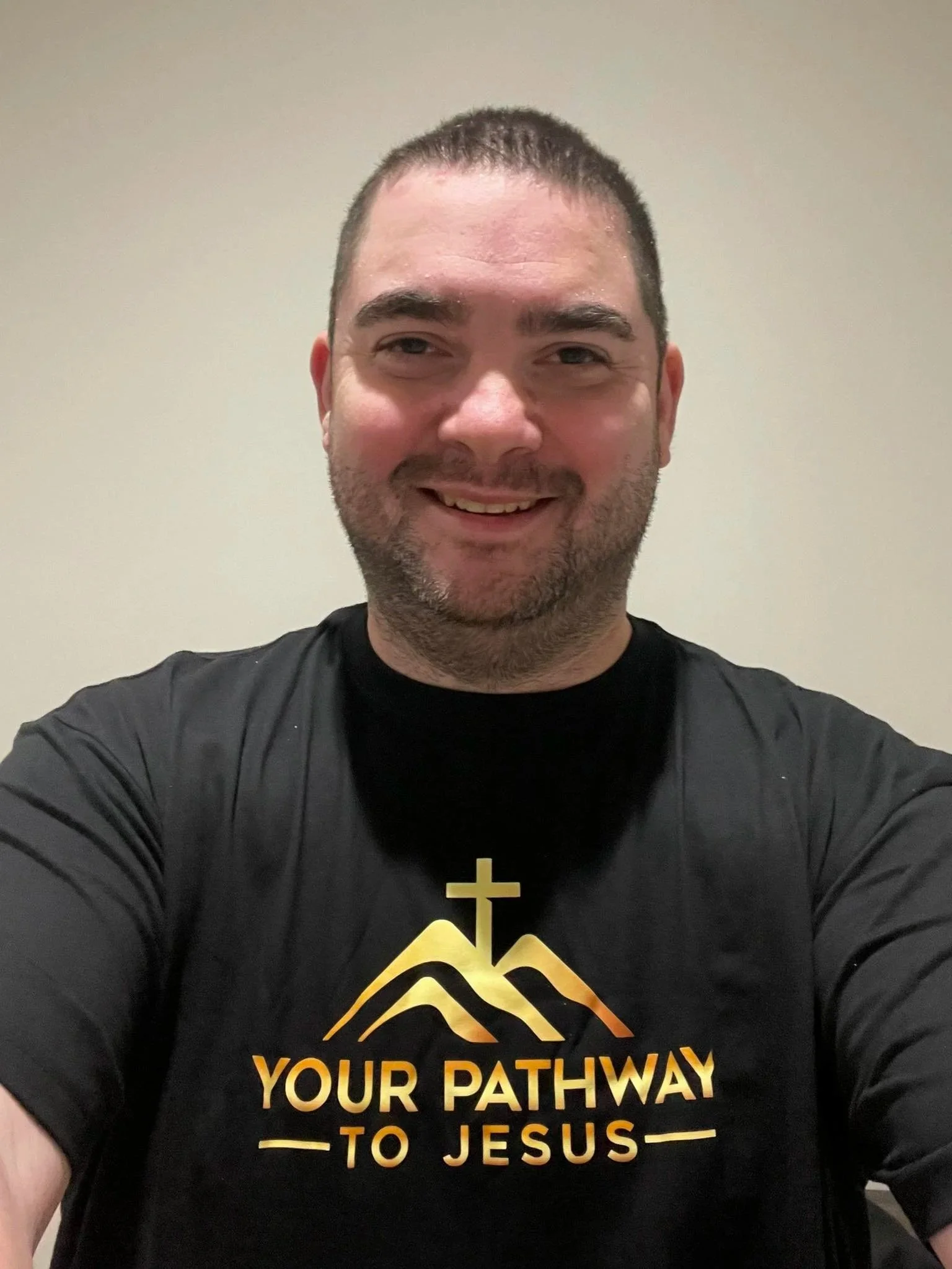 A man with short dark hair and a beard, smiling for a selfie, wearing a black T-shirt with a mountain, cross, and the words 'Your Pathway to Jesus' in gold lettering.