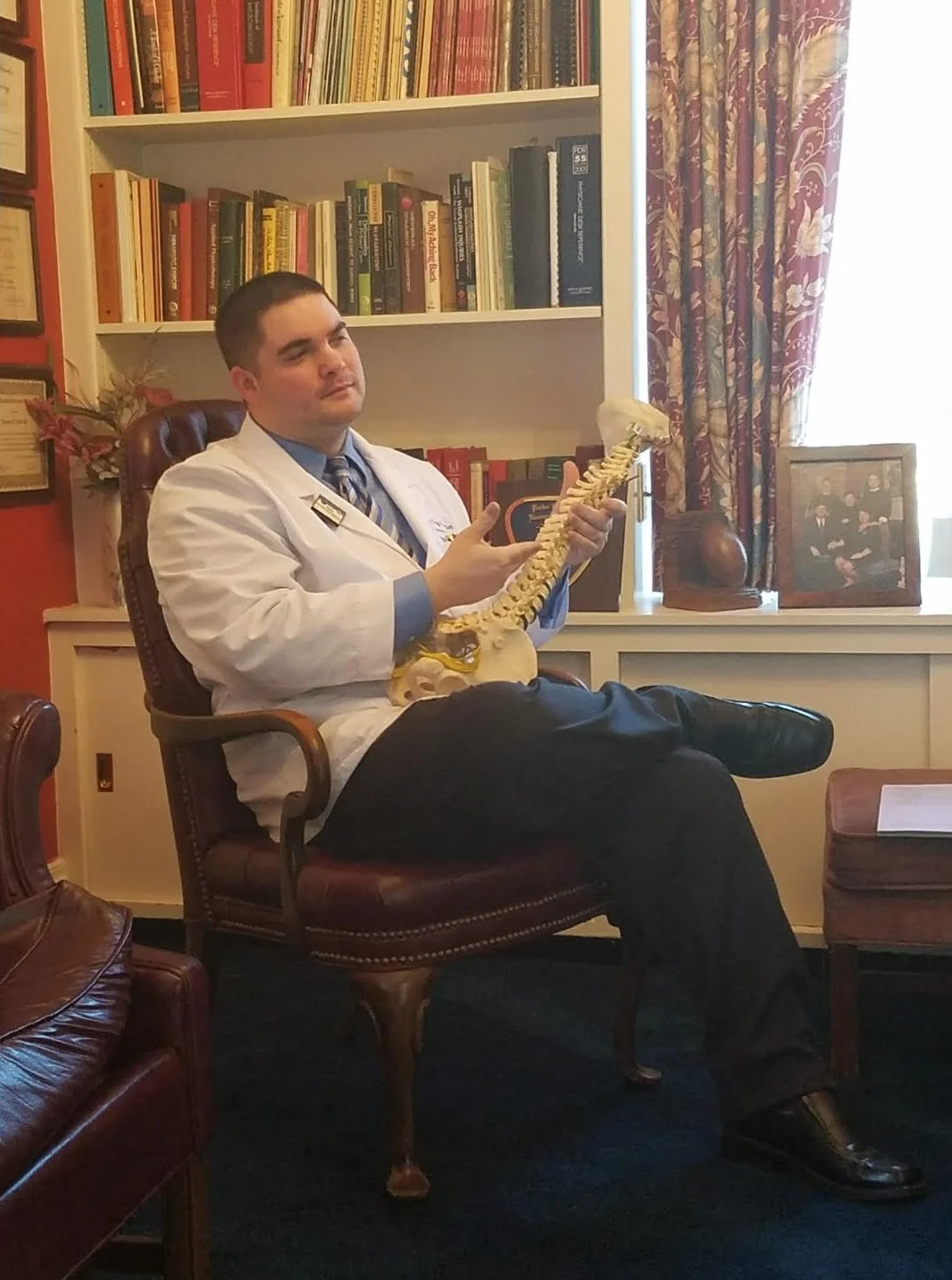 A man in a white coat sitting in a library, holding a model of a human spine and skull, with bookshelves and framed photographs in the background.