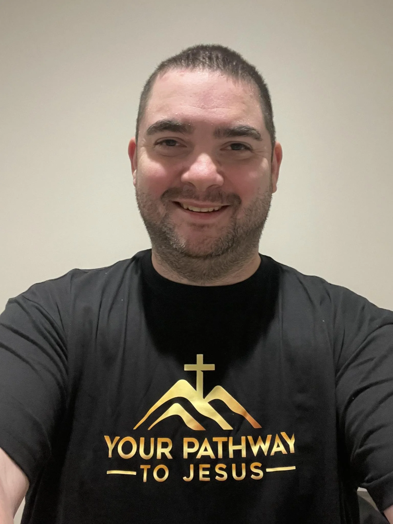 A man taking a selfie wearing a black t-shirt with a religious design and text that reads "Your Pathway to Jesus" against a plain beige wall background.