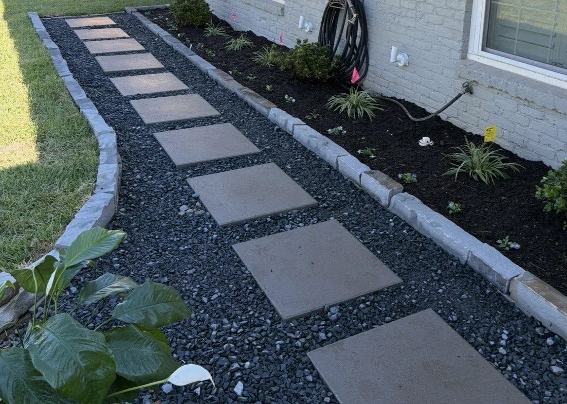 Landscaped side yard with square stepping stones set in black gravel beside a white brick house and planted flower bed