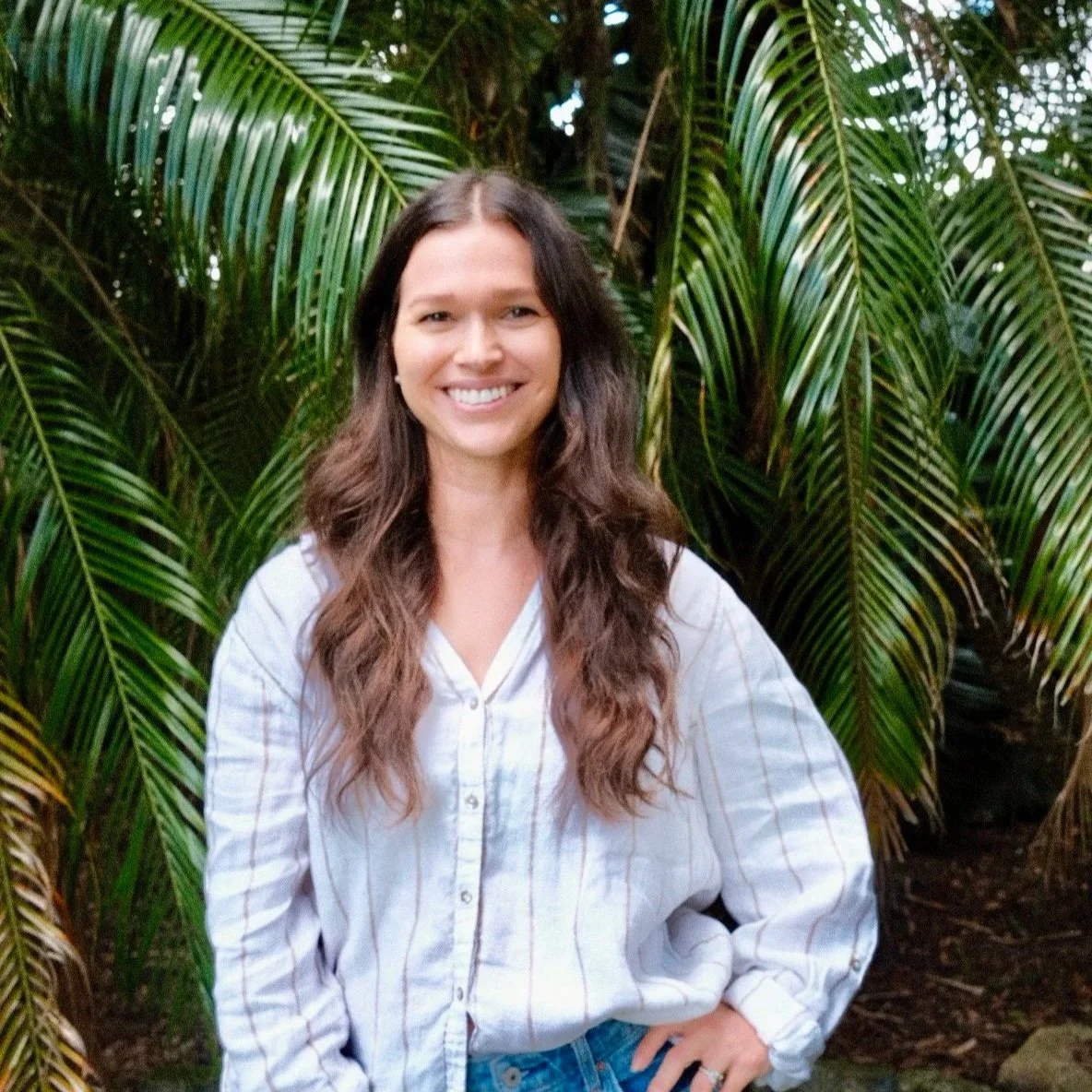 A woman with long wavy brown hair smiling and standing outdoors in front of lush green palm leaves.