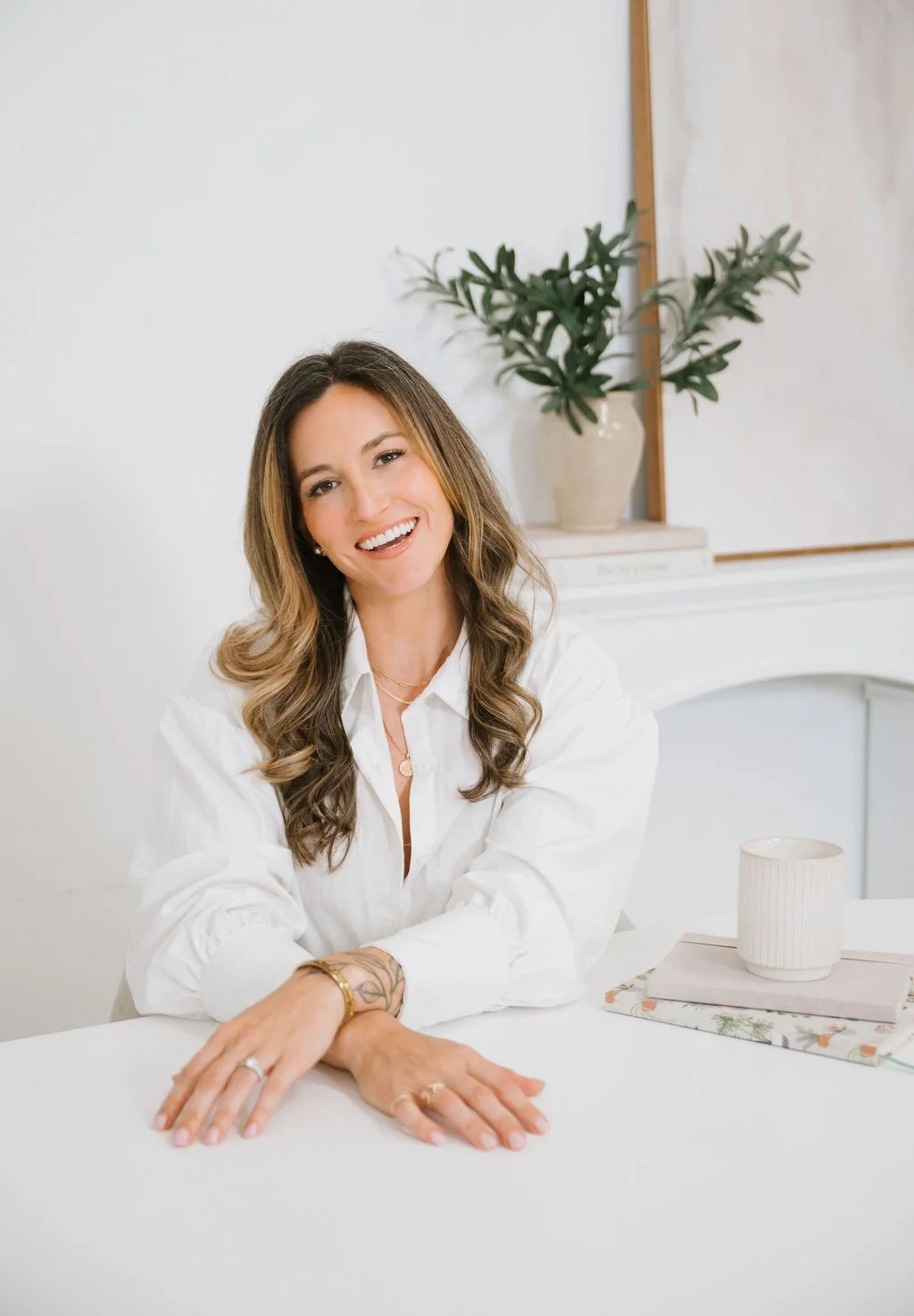 A smiling woman with long wavy brown hair, wearing a white shirt, sitting at a white table with a notebook, a mug, and a plant in the background.