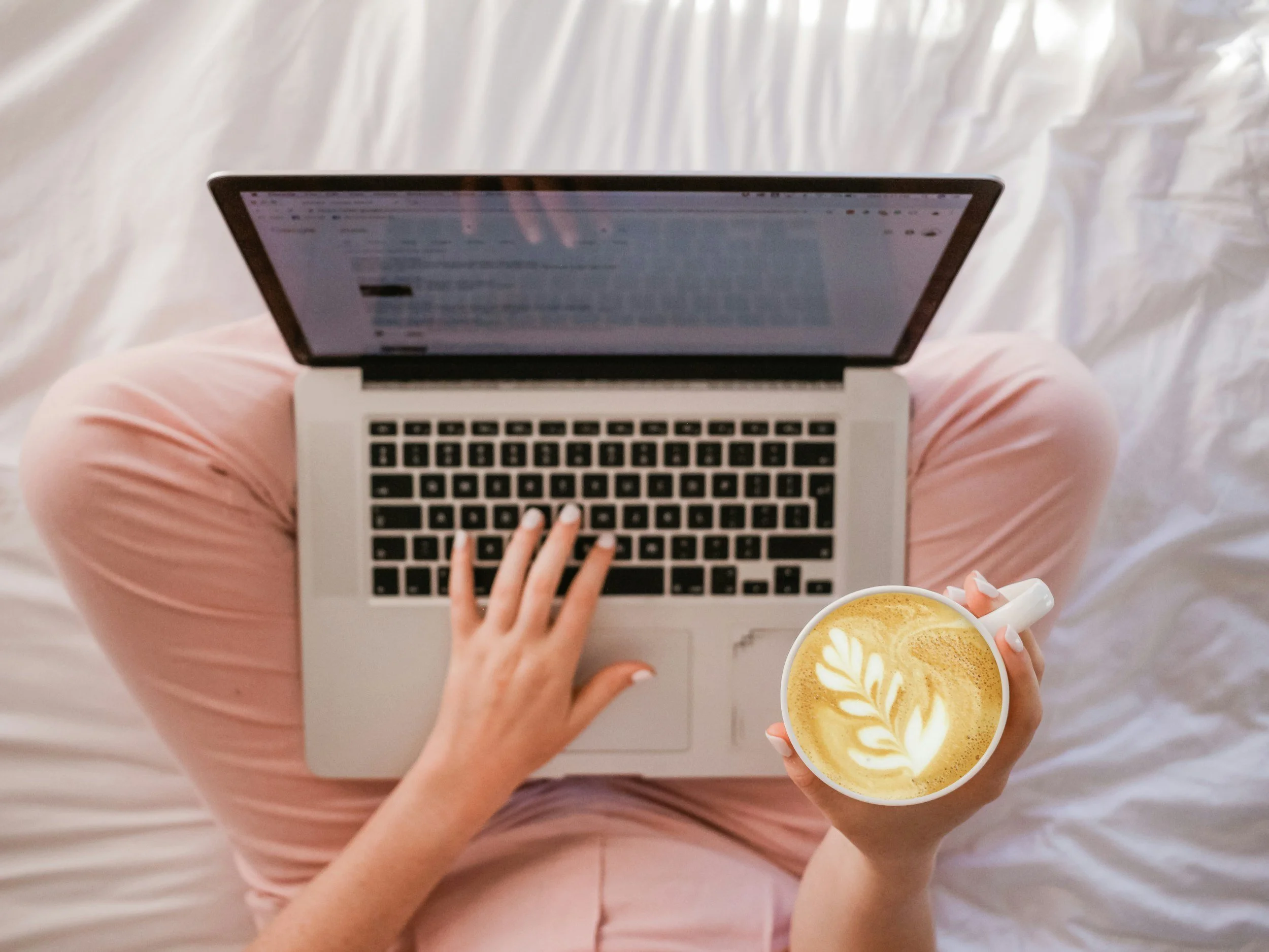 Person sitting on a bed with pink pants, using a silver laptop, holding a cup of coffee with latte art.