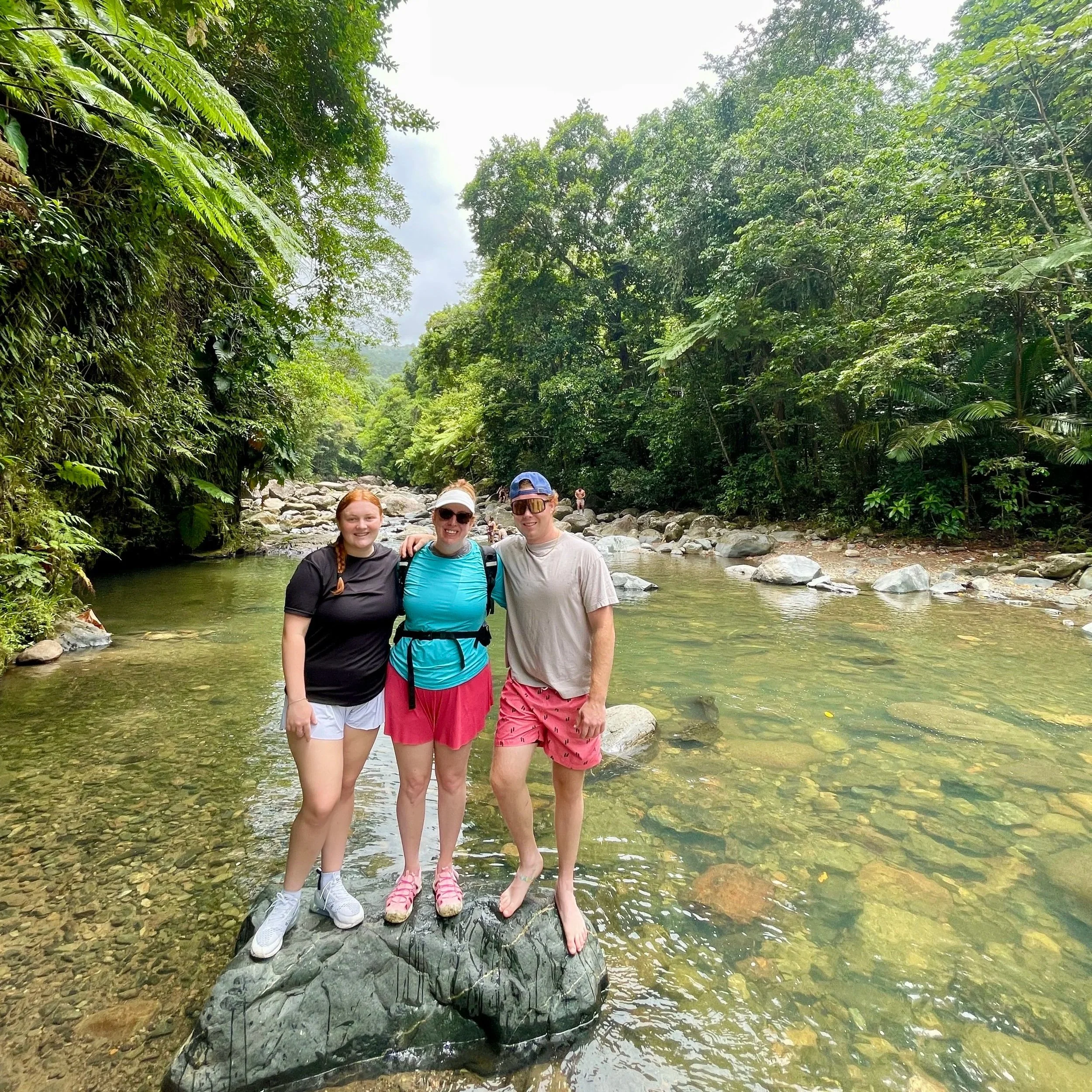 A woman in a black graduation gown and cap walking on an outdoor corridor with glass windows and stone columns.
