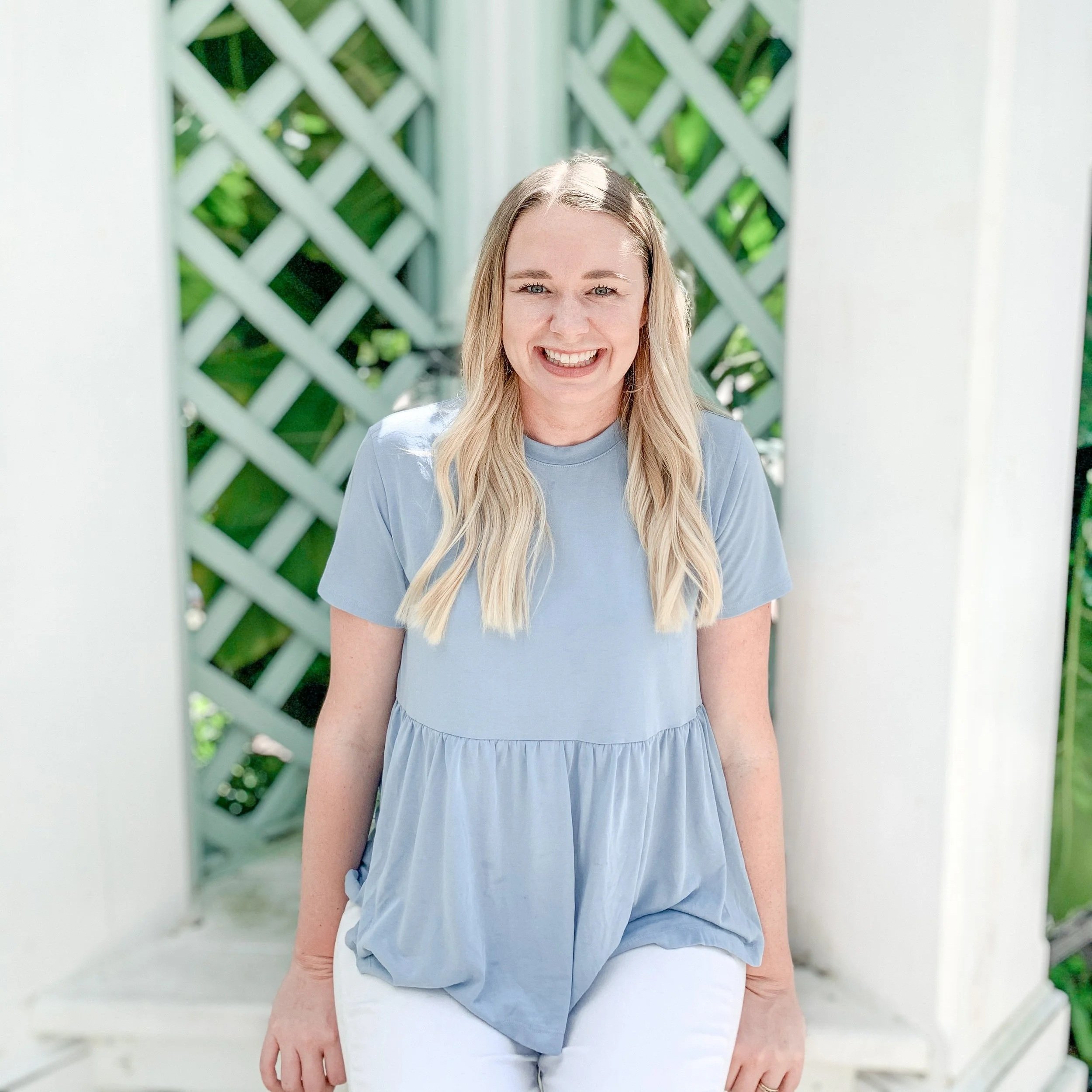 A woman with blonde hair wearing a light blue top and white pants, standing outdoors in front of white lattice fencing with green plants behind.