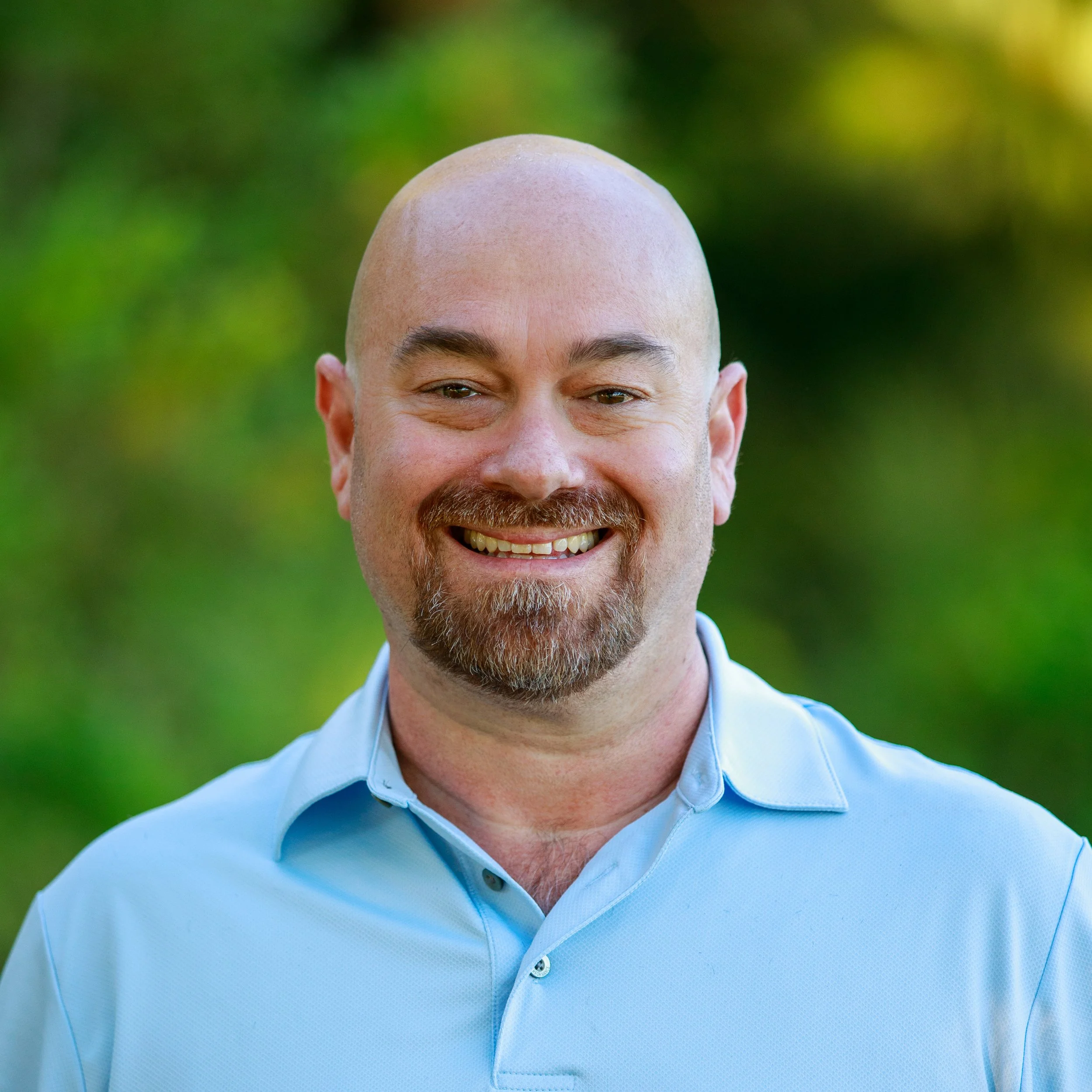 Close-up of a man smiling, with a shiny bald head, gray beard, wearing a light blue collared shirt, outdoors with green blurred background.
