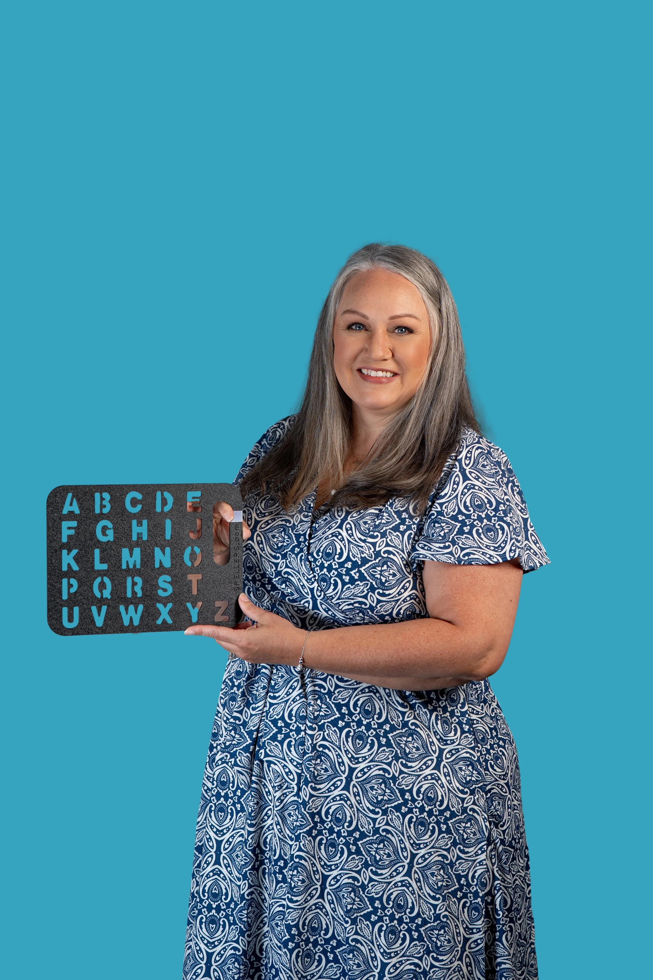 A middle-aged woman with long gray hair smiling and holding an eye chart against a plain blue background.