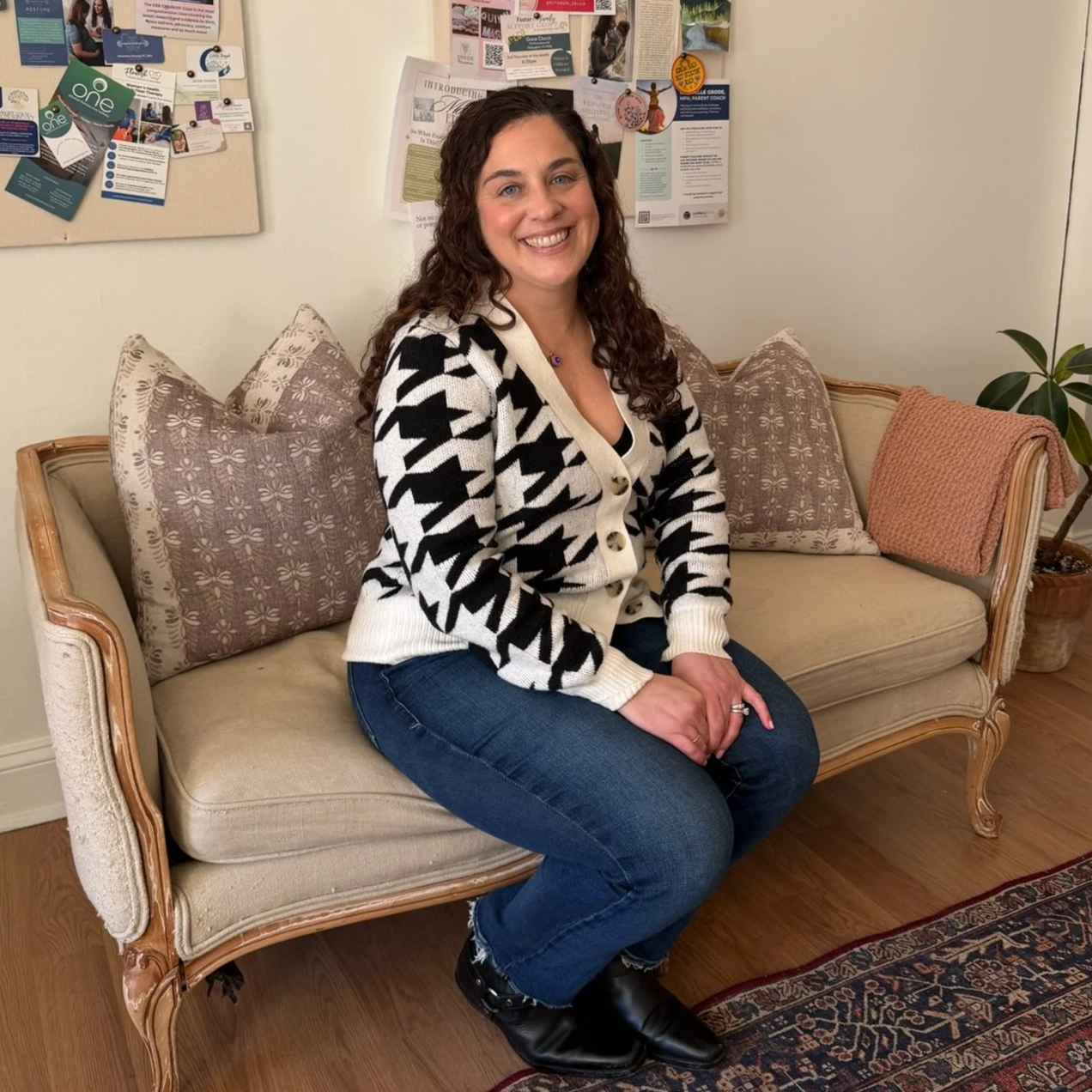A woman with long, curly brown hair, smiling, sitting on a beige vintage-style sofa with patterned pillows, in a room with a bulletin board and potted plant in the background.