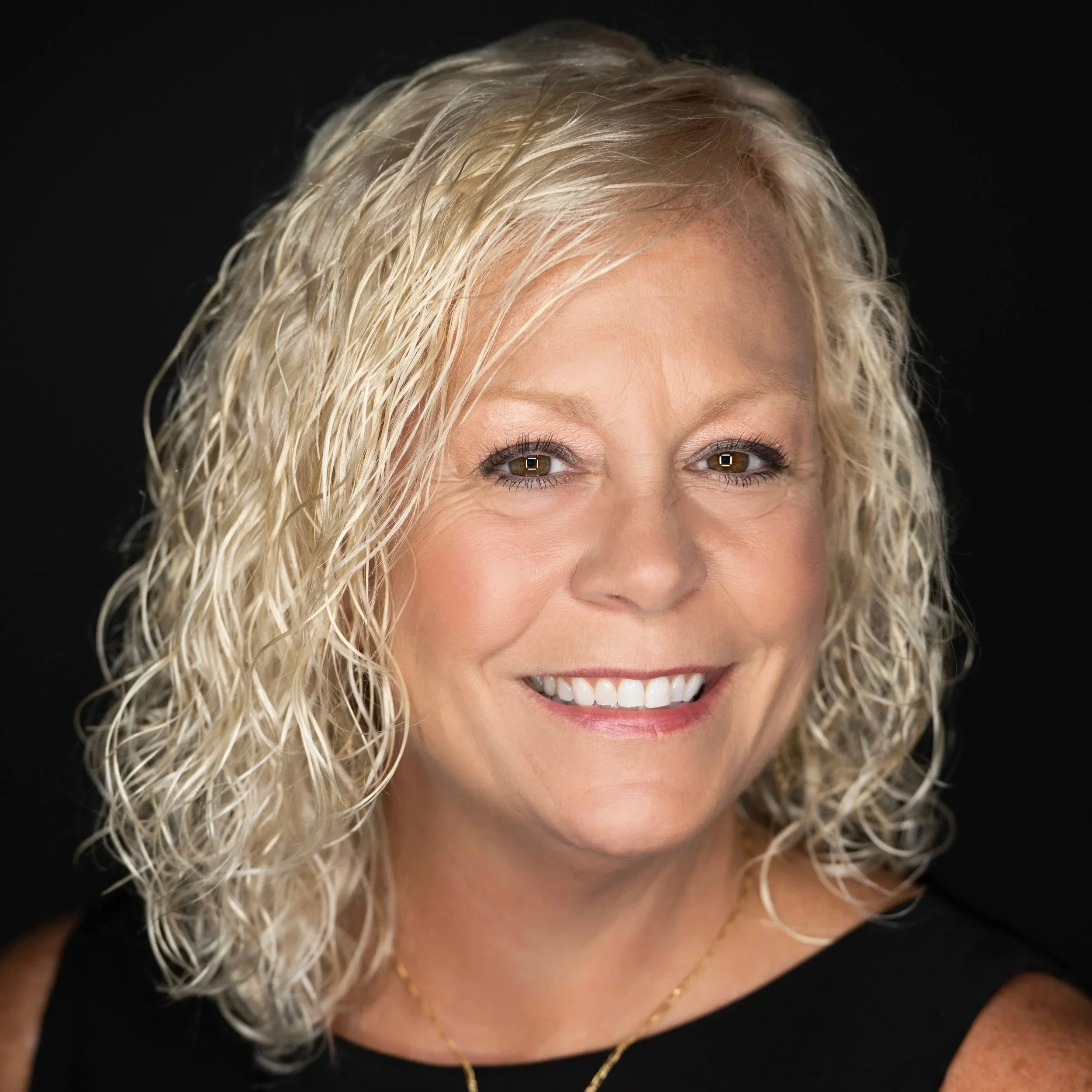 Close-up of a woman with blonde, curly hair smiling, wearing a black top and gold jewelry, against a black background.