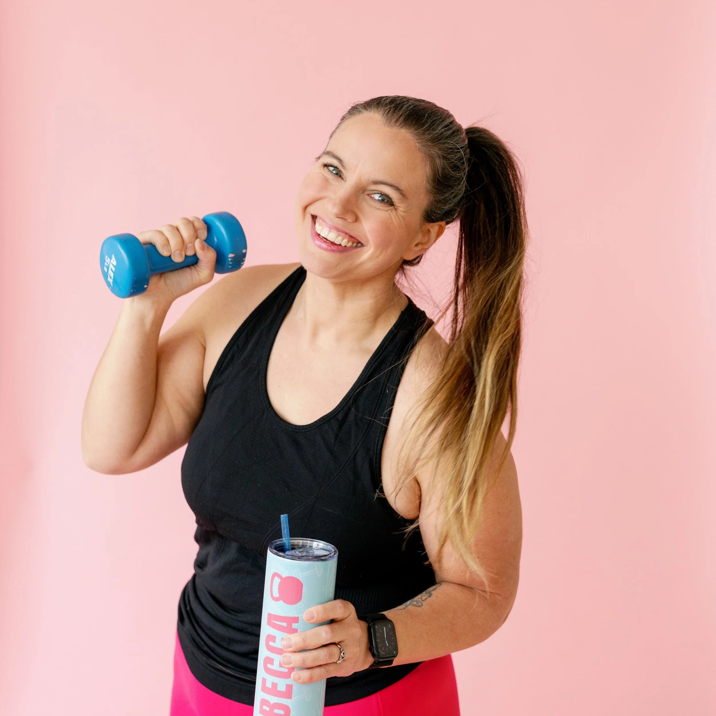 A woman smiling and holding a light blue cup with pink lettering, wearing a black tank top, pink leggings, and a smartwatch, standing against a pink background.