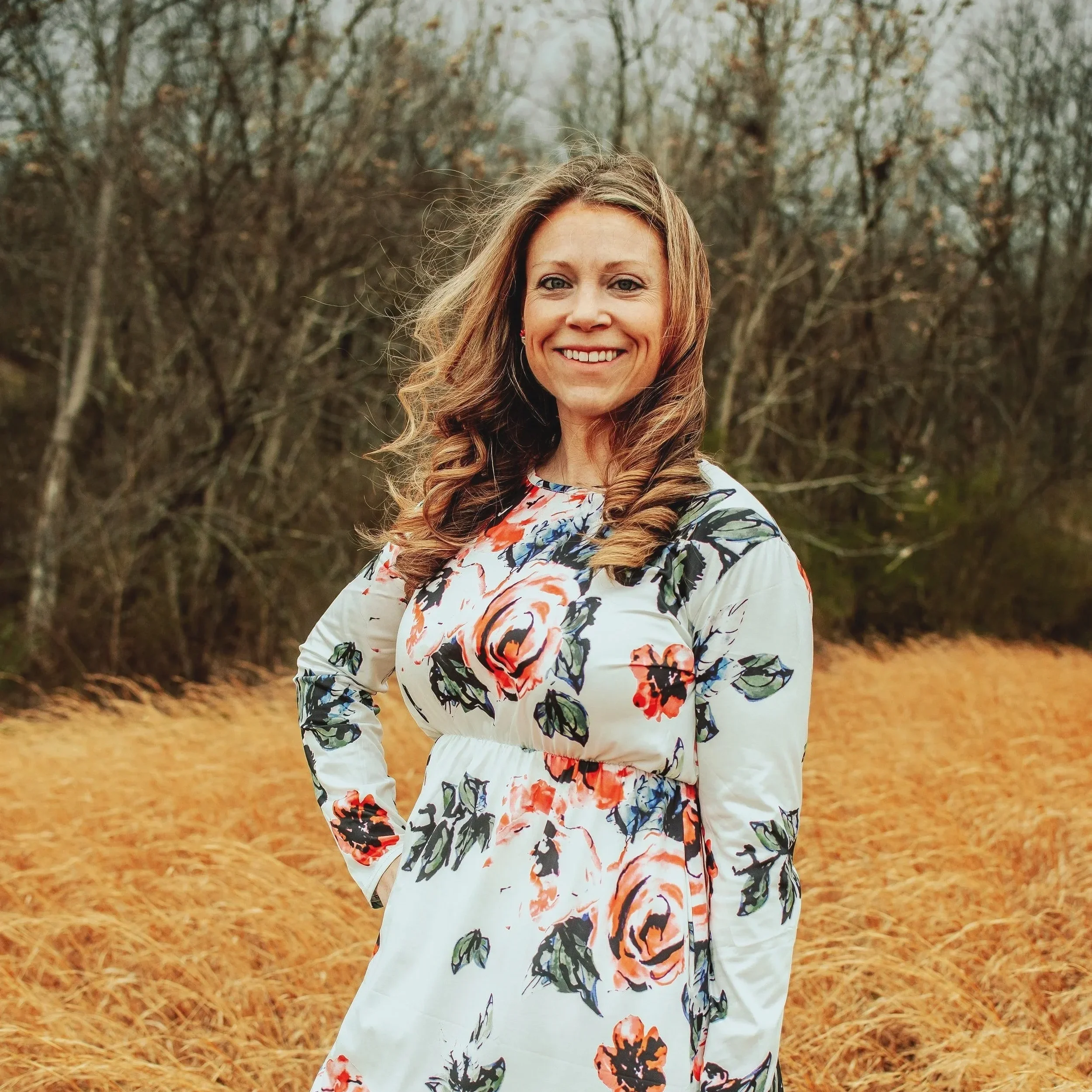 A woman with curly light brown hair smiling outdoors on a fall day, wearing a floral dress with long sleeves, standing in front of leafless trees and a bed of fallen leaves.