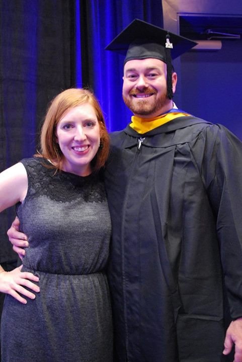 A woman with red hair and a man in a graduation cap and gown smiling together at a graduation ceremony.