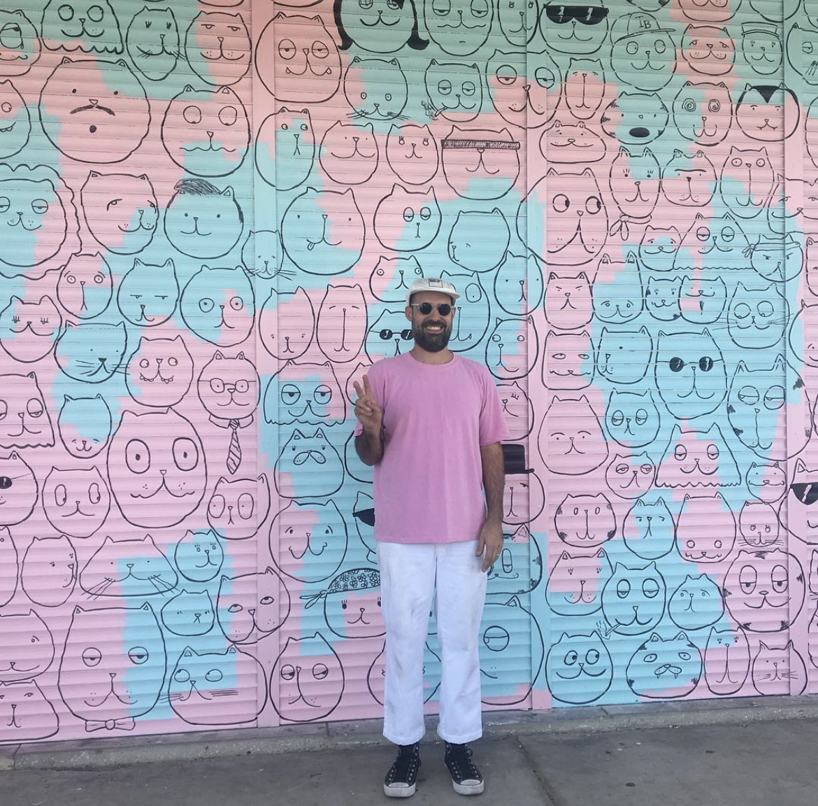 A man in a pink T-shirt, white pants, and sunglasses standing in front of a colorful mural wall decorated with many simple, cartoonish black line drawings of cats in various expressions and accessories, making a peace sign with his right hand.