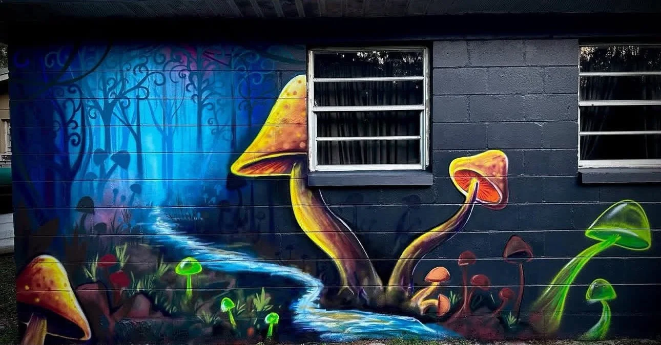 Colorful mural of a whimsical forest scene with a glowing stream, large mushrooms, and trees on a dark wall, featuring two window openings.