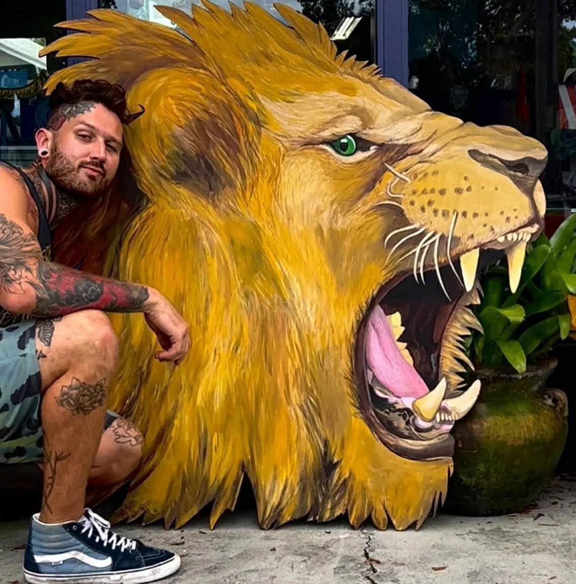 A man with tattoos squats next to a large wooden cutout of a roaring lion.
