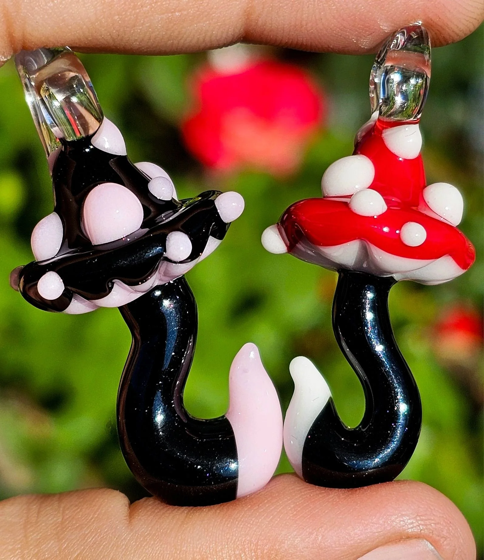 Two glass mushroom-shaped ornaments, one red with white spots and one black with white spots, being held between a person's fingers with a garden background.