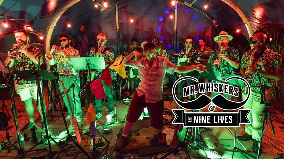 A lively band called 'Mr. Whiskers & The Nine Lives' playing tropical-themed music. The band members are dressed in colorful Hawaiian shirts, some wearing sunglasses, and are playing various instruments including saxophones and a keyboard. The stage is decorated with string lights and flags, creating a festive atmosphere.