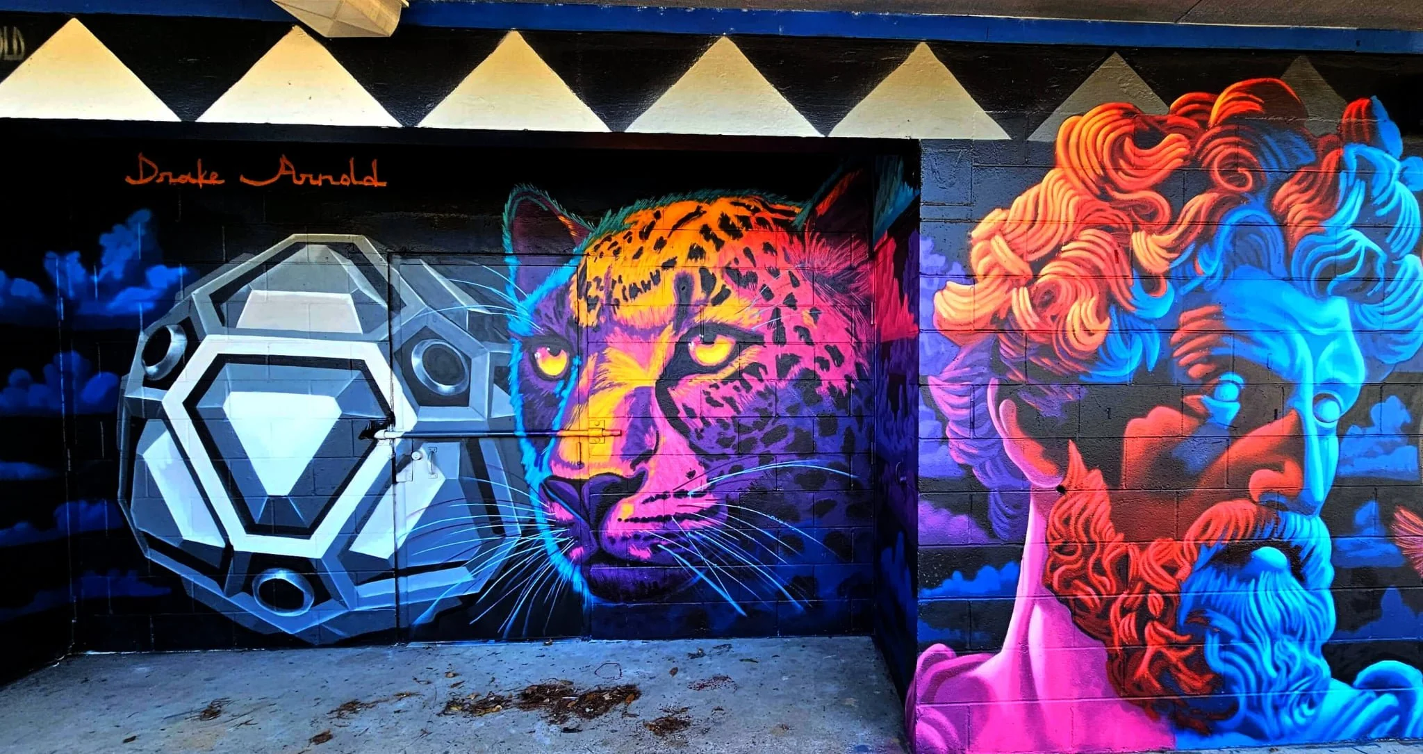 Colorful mural with a geometric sphere on the left, a neon-lit leopard's face in the center, and a classical sculpture of a man's head on the right, all painted on a brick wall.