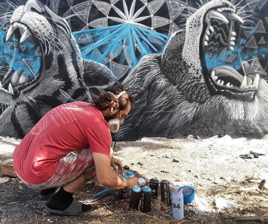 A person spray painting a mural of two roaring lions on a wall, with spray paint cans and tools on the ground.