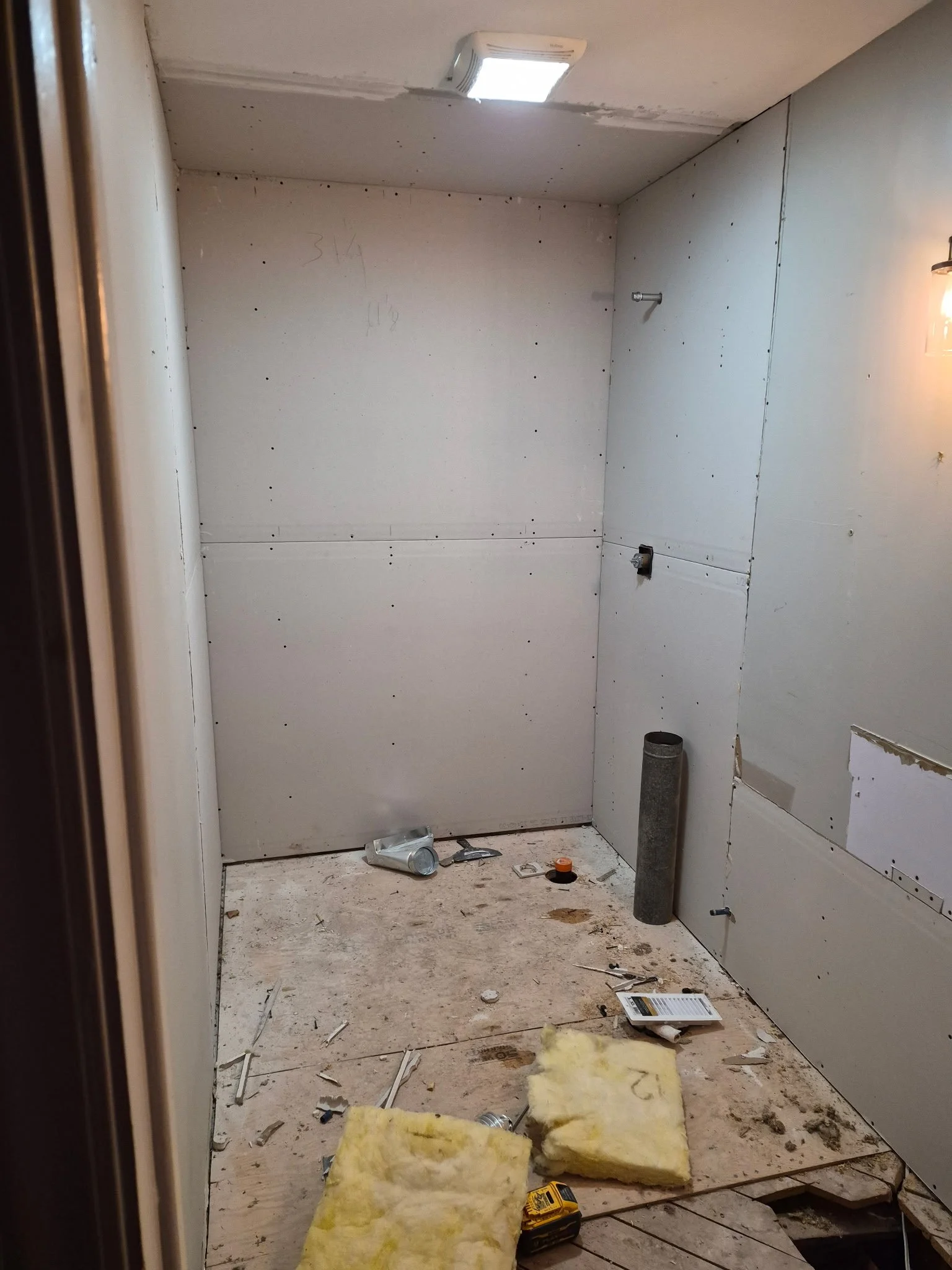 Small, unfinished room with drywall walls, construction tools and debris on the floor, yellow insulation, a roll of insulation, a flashlight, and a ceiling light fixture.