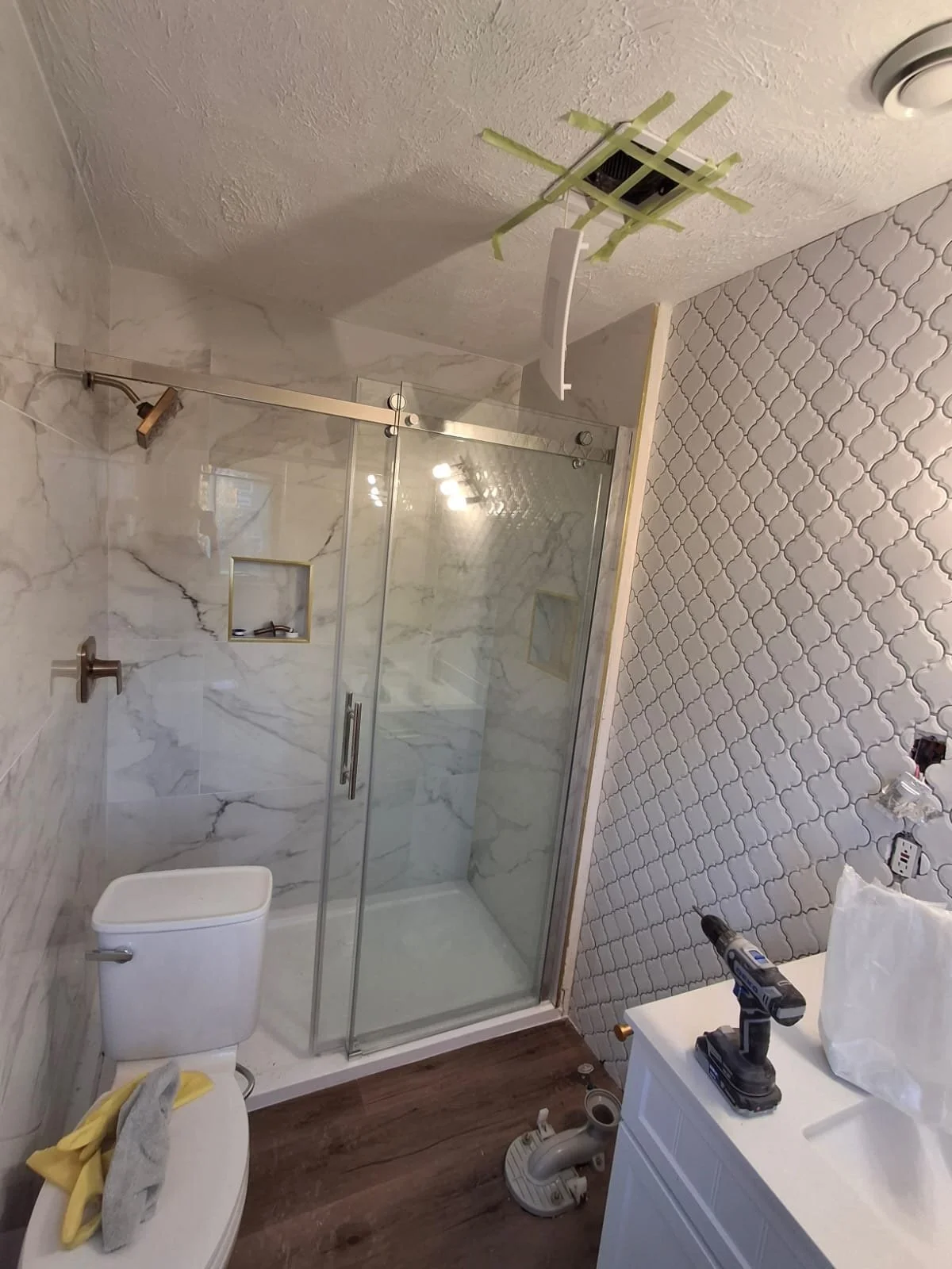 Bathroom in the process of being renovated; includes a shower with glass door, toilet, vanity with tools, and wall with patterned tile.