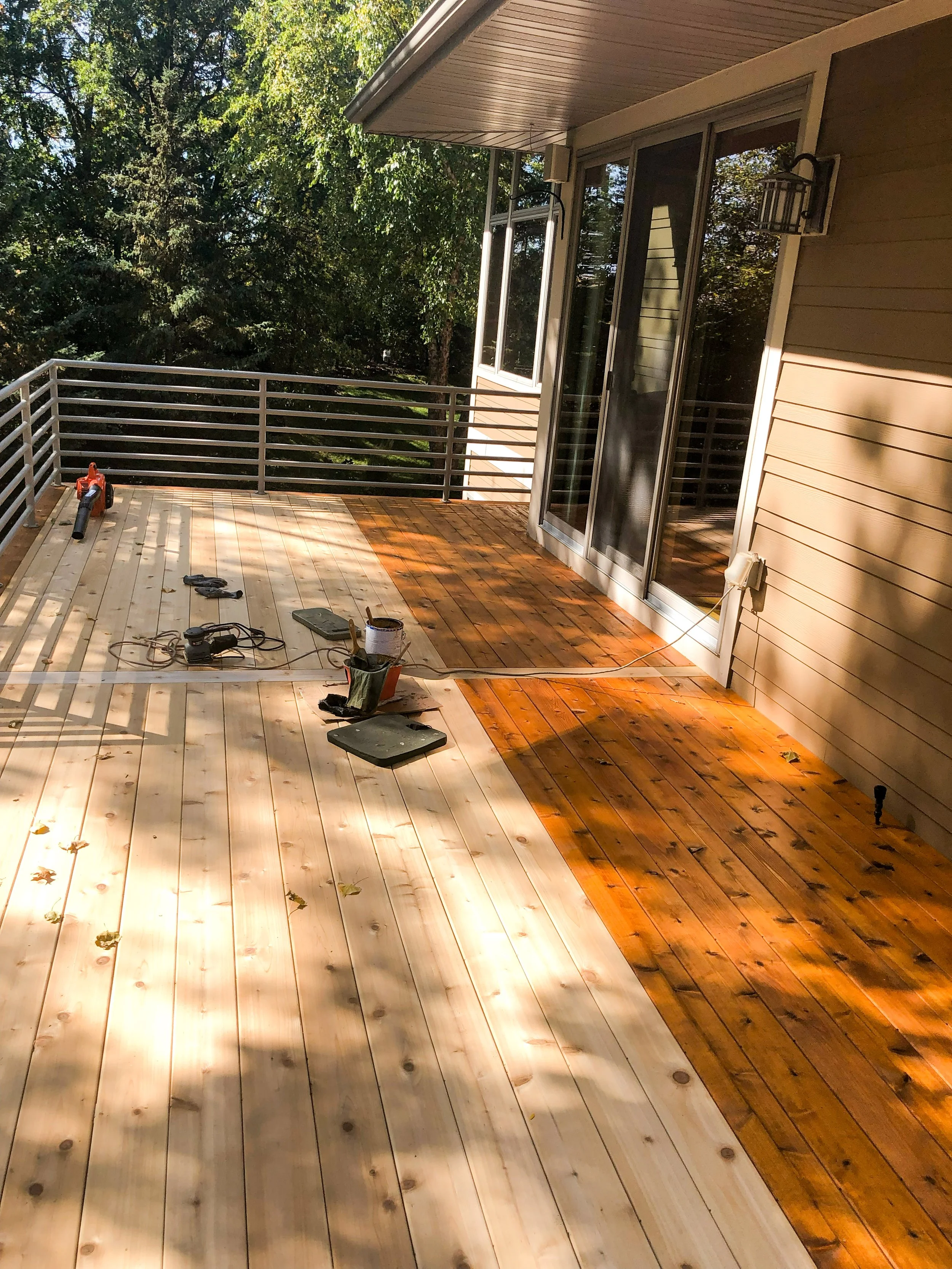 A deck under construction with some wooden planks partially stained in a darker color, tools and supplies scattered around, and a view of trees and greenery in the background.