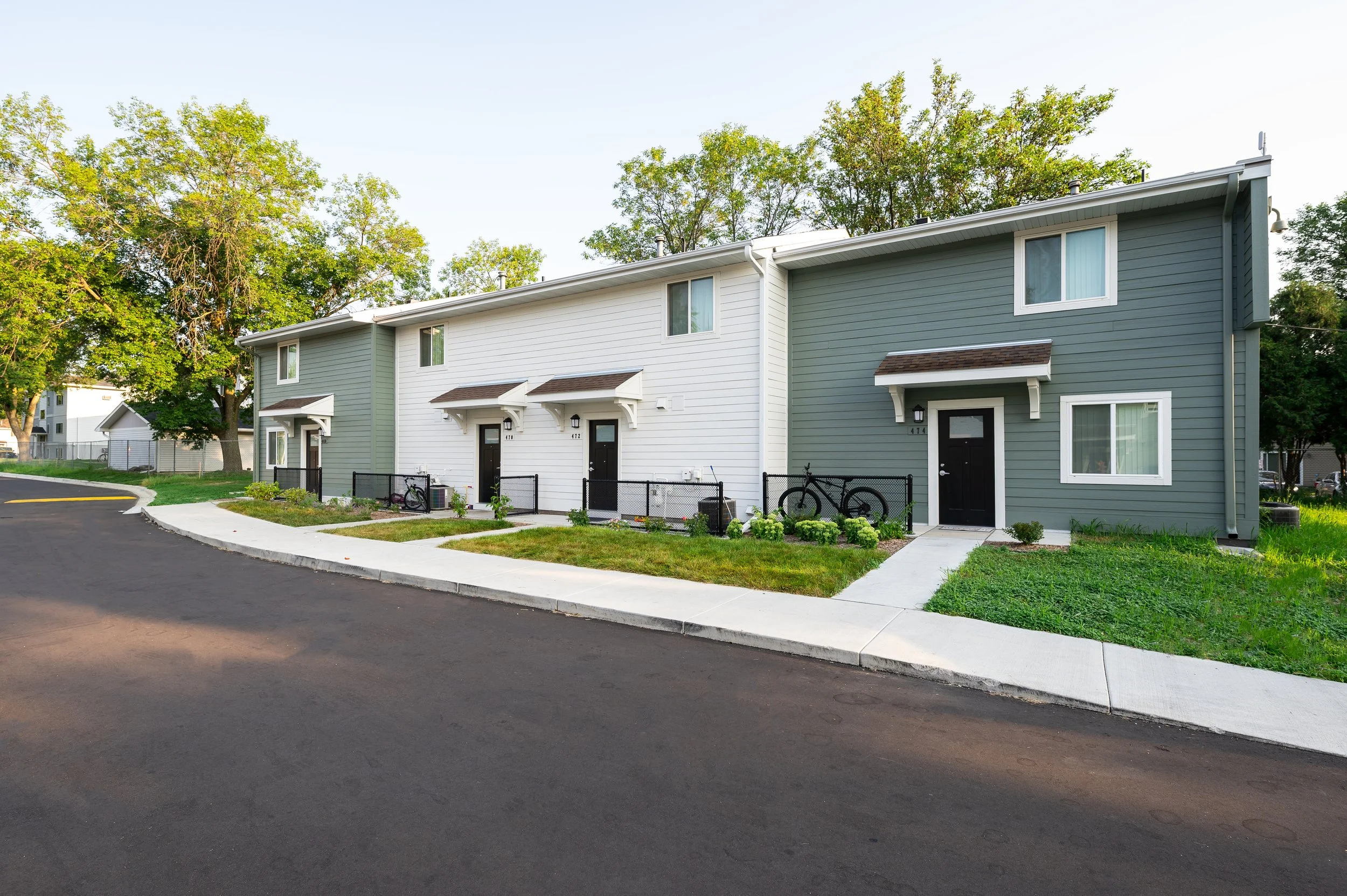 A row of modern townhouses with black doors, small front gardens, and bicycles parked outside, set against a backdrop of trees and a clear sky.