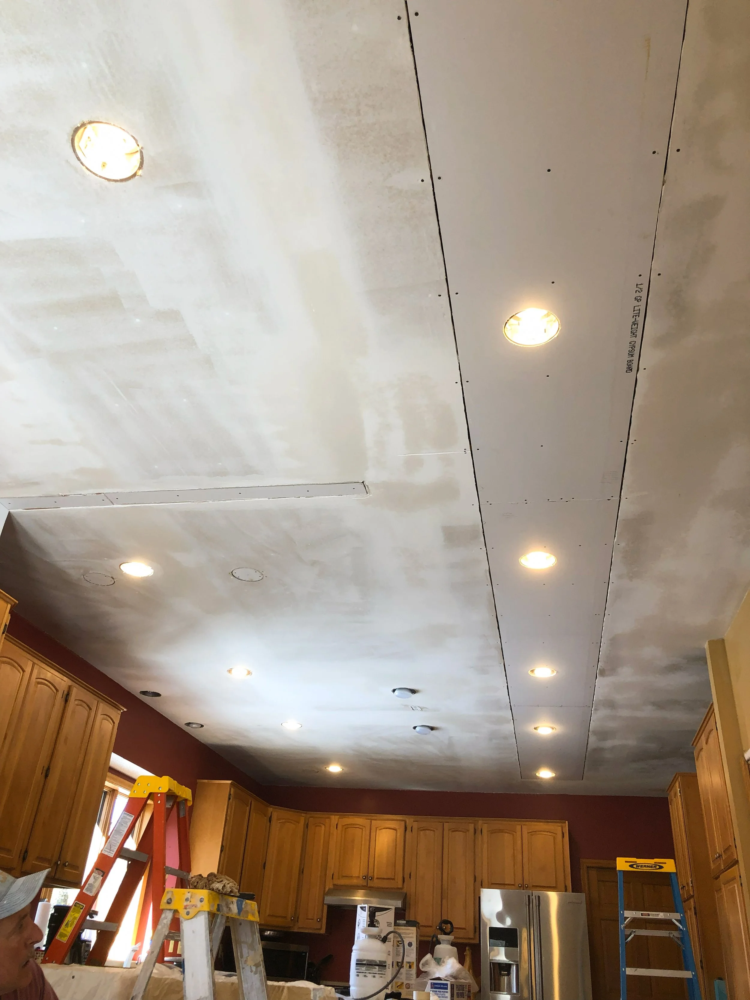 Kitchen ceiling undergoing renovation, with recessed lighting installed, and construction tools like ladders and supplies visible.
