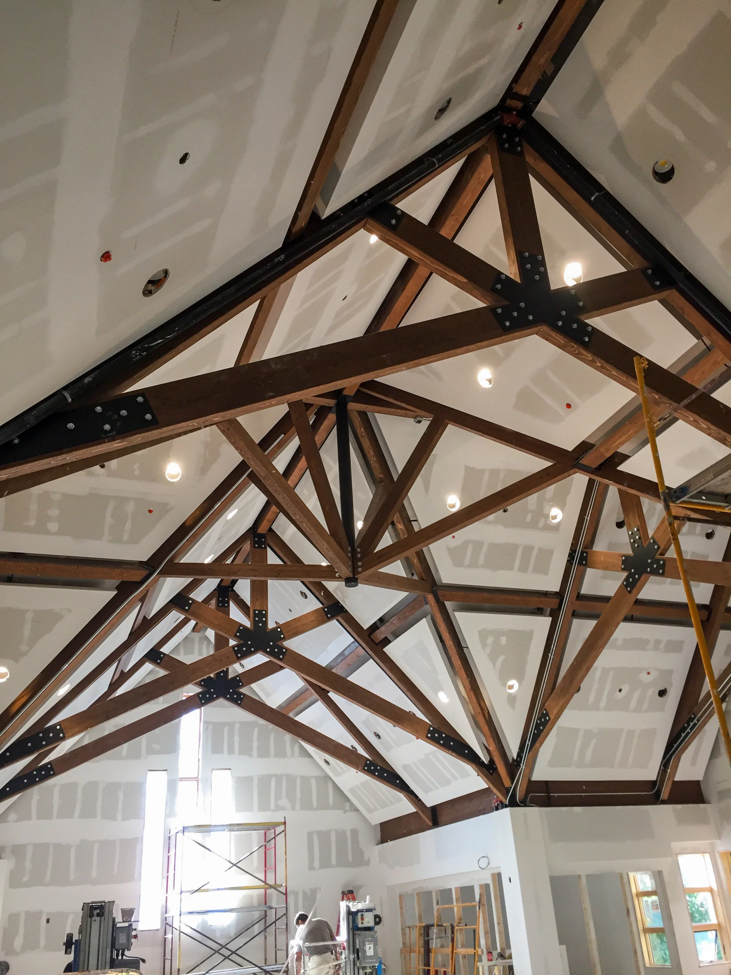 Construction site showing an unfinished ceiling with exposed wooden beams and black metal connectors, with some insulation and drywall installed.