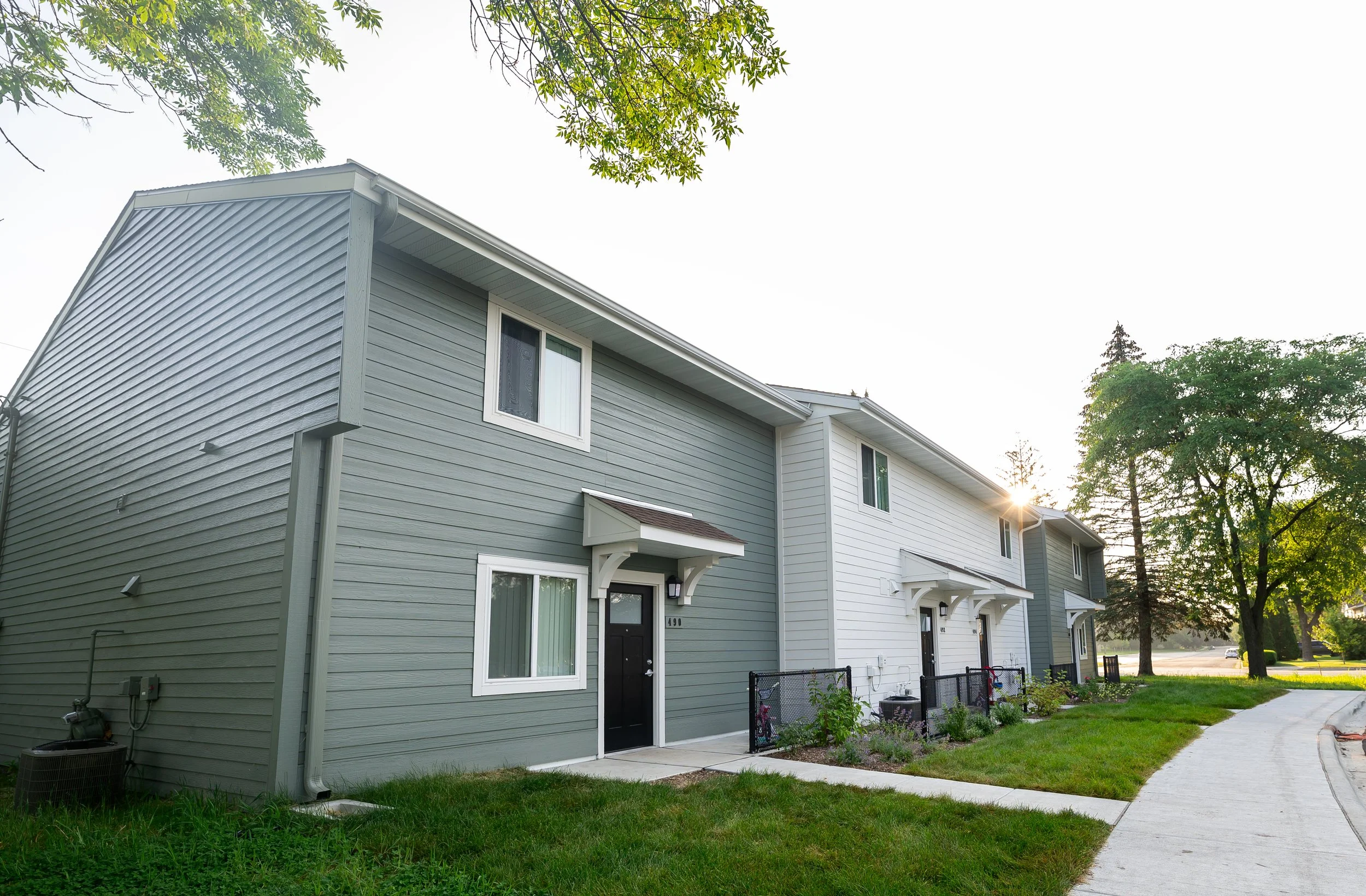 A row of modern townhouses with gray and white siding, small front porches with awnings, overlooking a sidewalk with grass and trees in the background, during sunset.