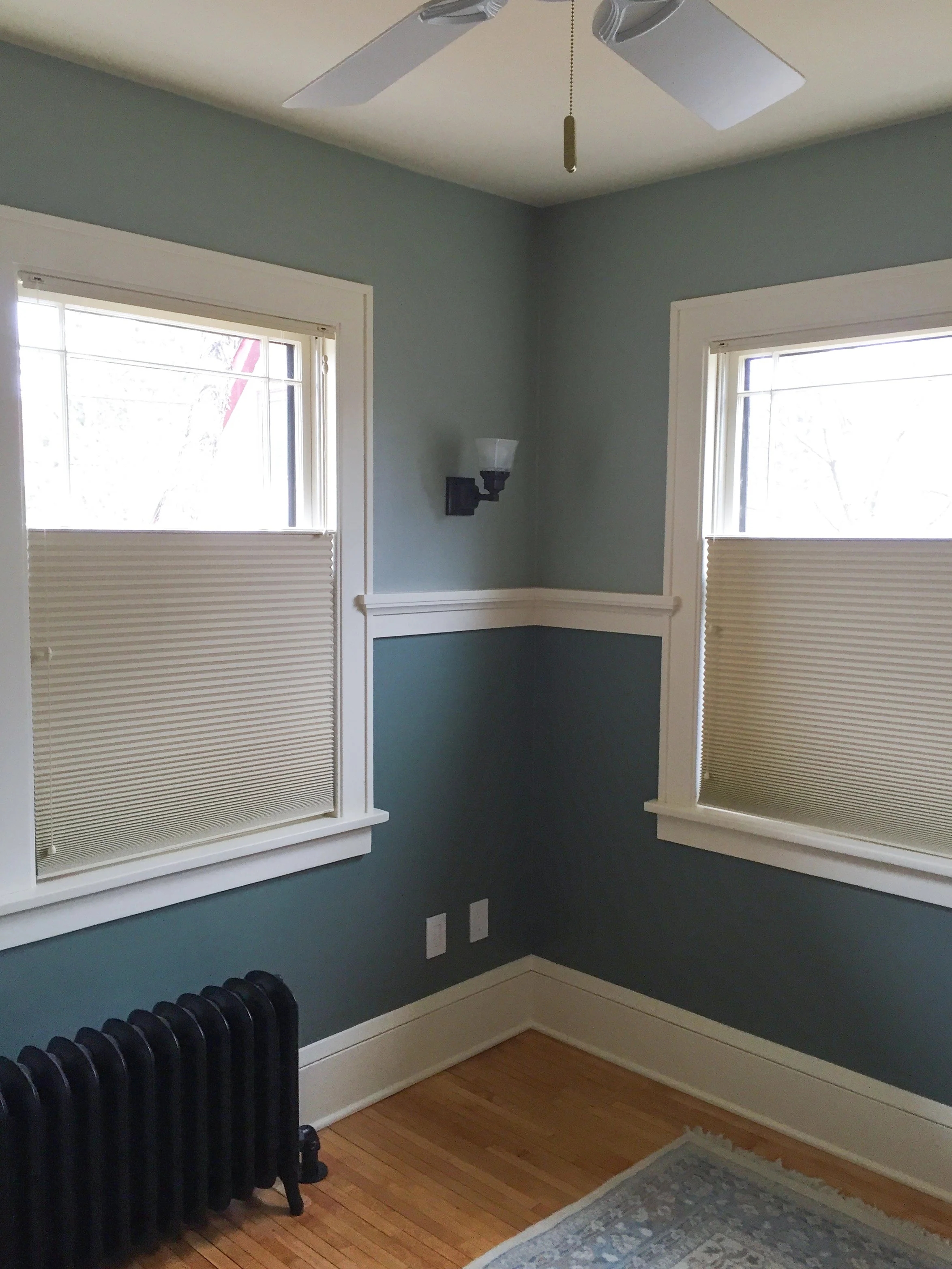 An empty room with two windows covered by beige pleated shades, a black radiator against the wall, a wall sconce light, white trim, and a ceiling fan with white blades, on a hardwood floor.