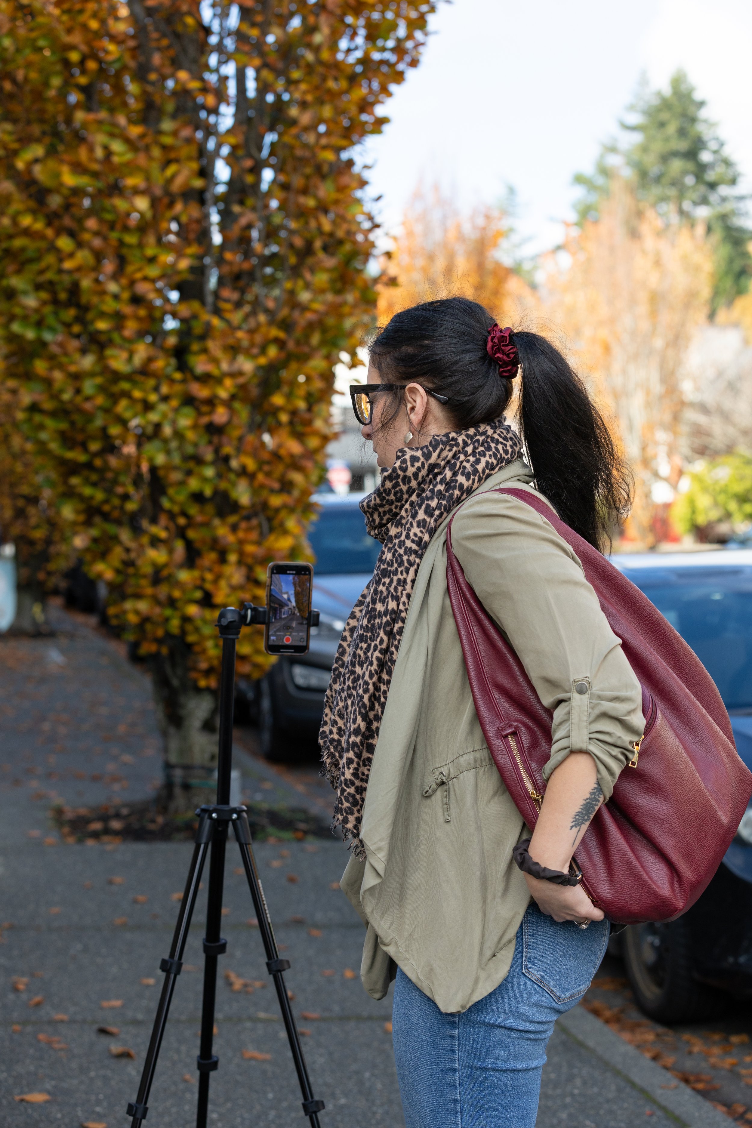 Woman with dark hair tied in a ponytail, wearing glasses, a leopard print scarf, a beige jacket, and jeans, standing outdoors in autumn, filming with a smartphone on a tripod.